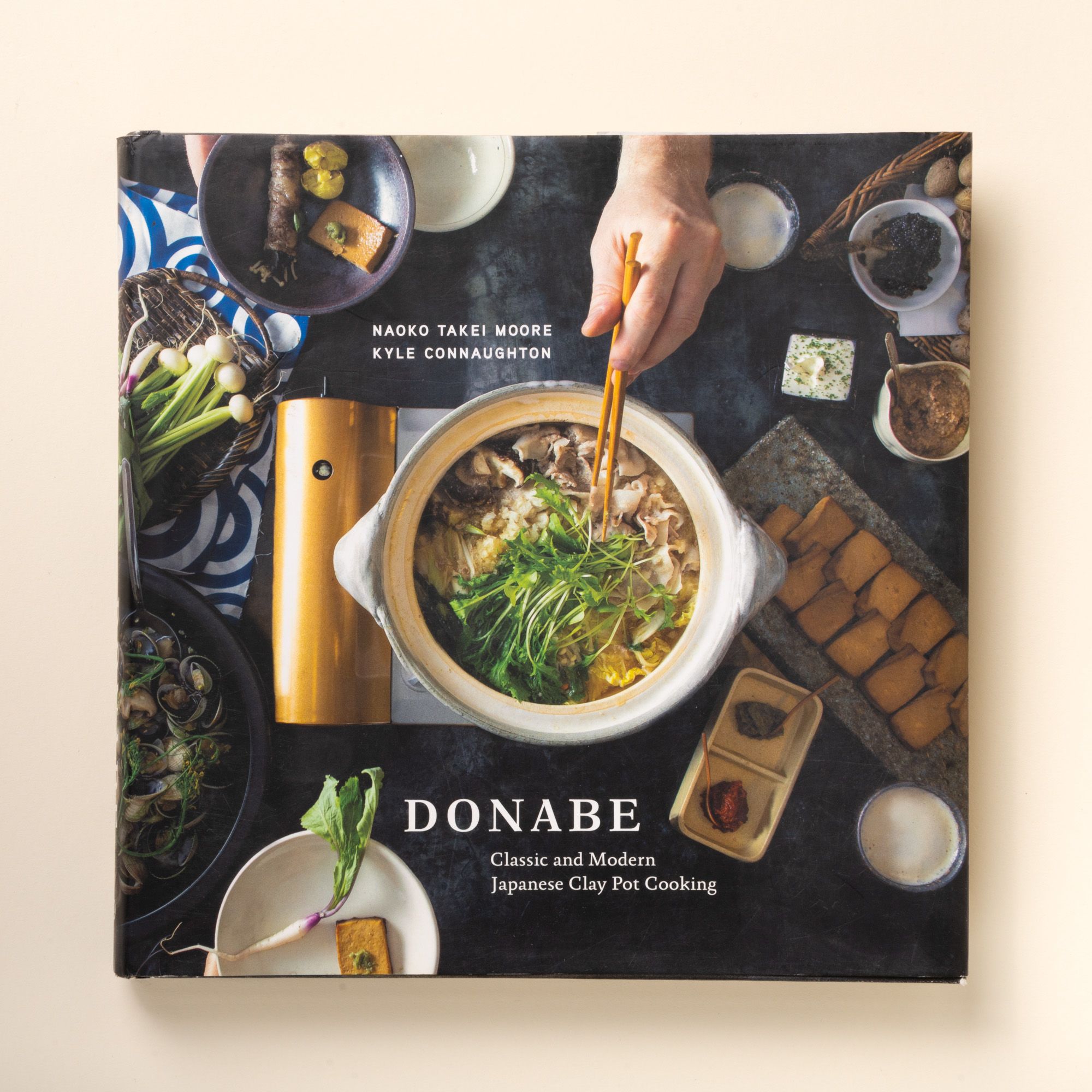 Front cover of "Donabe," showing a clay pot filled with soup and greens surrounded by bowls and ingredients, a hand reaching in with chopsticks