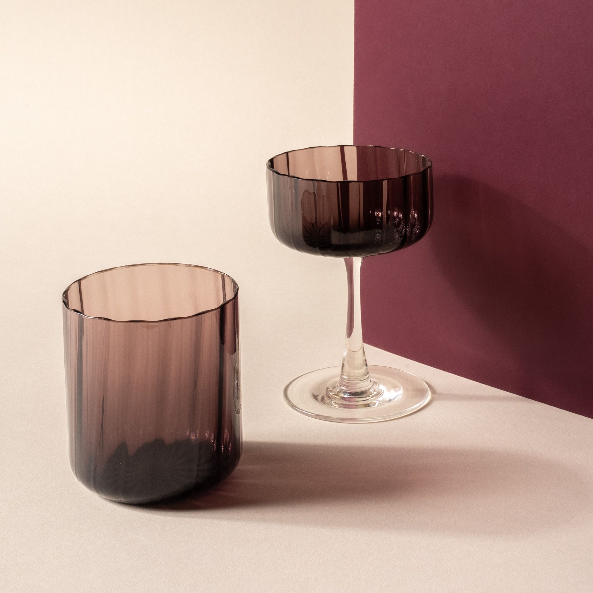 A fluted walled tumbler and coupe glass with a fluted bowl that is a grey purple color with a clear stem, with a purple wall prop.