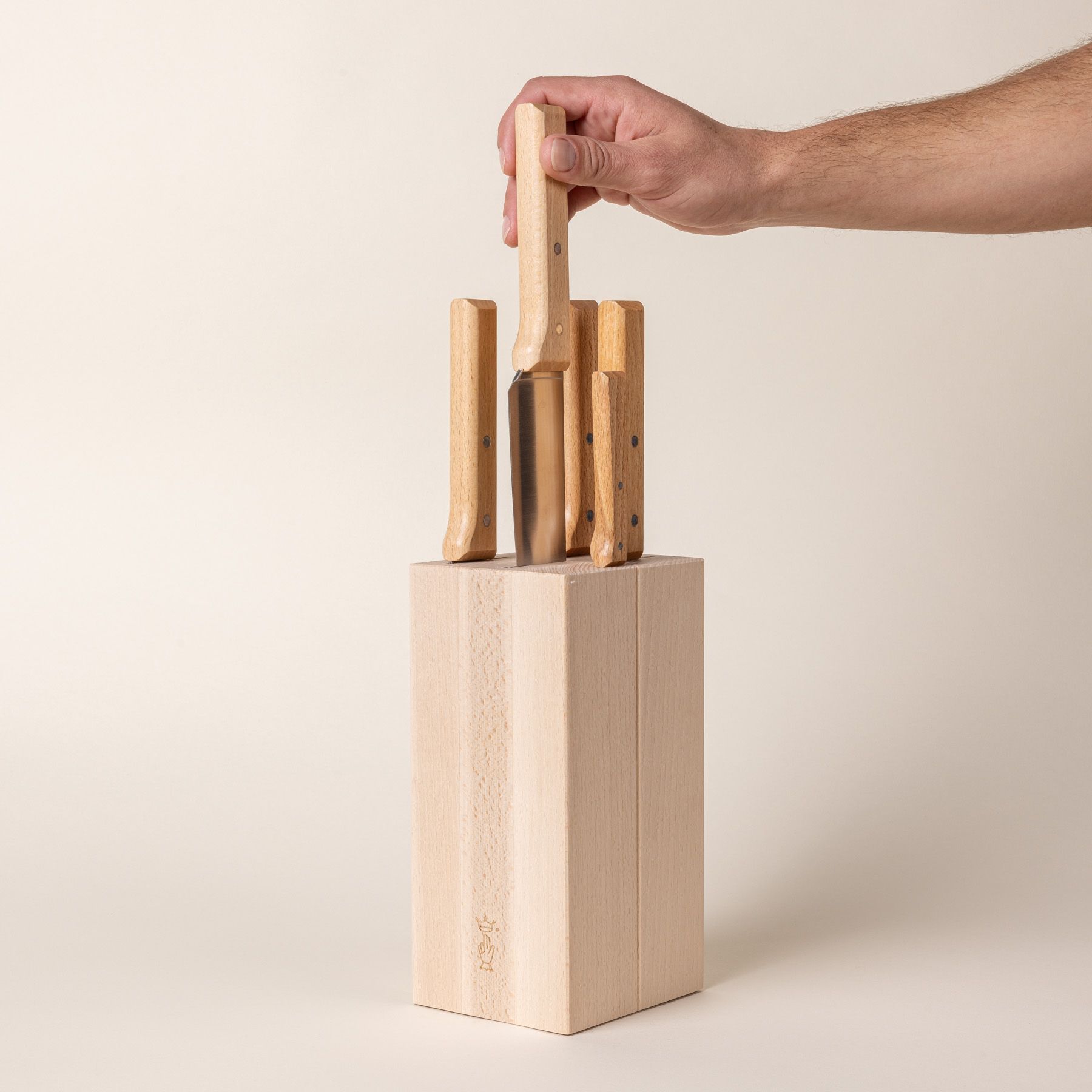 A tall rectangular wooden block with knives inside and the wooden handles sticking out at the top. A hand is grabbing one of the knives out of the block.