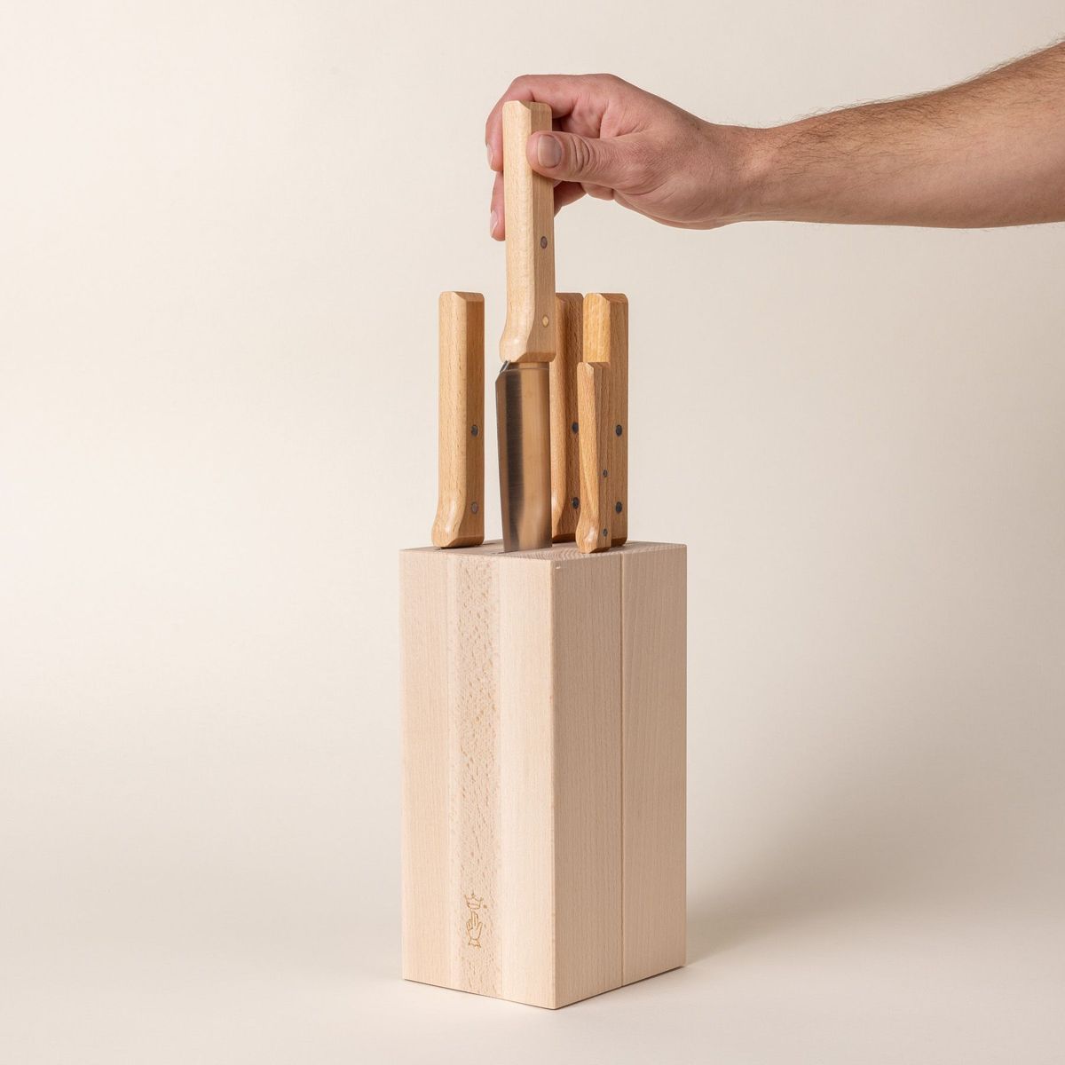 A tall rectangular wooden block with knives inside and the wooden handles sticking out at the top. A hand is grabbing one of the knives out of the block.