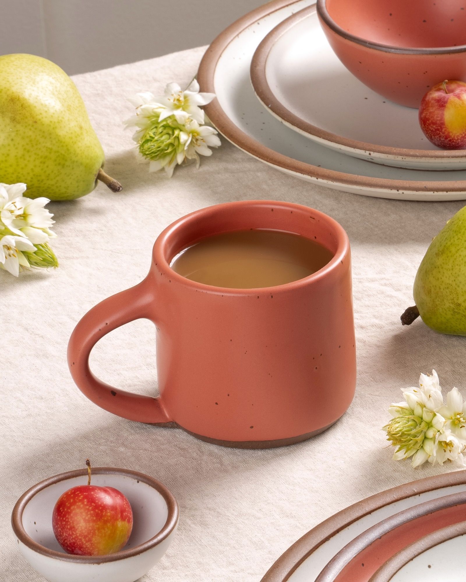 Coffee in a watermelon pink mug with pears, flowers, and stoneware plates on a neutral tablecloth.