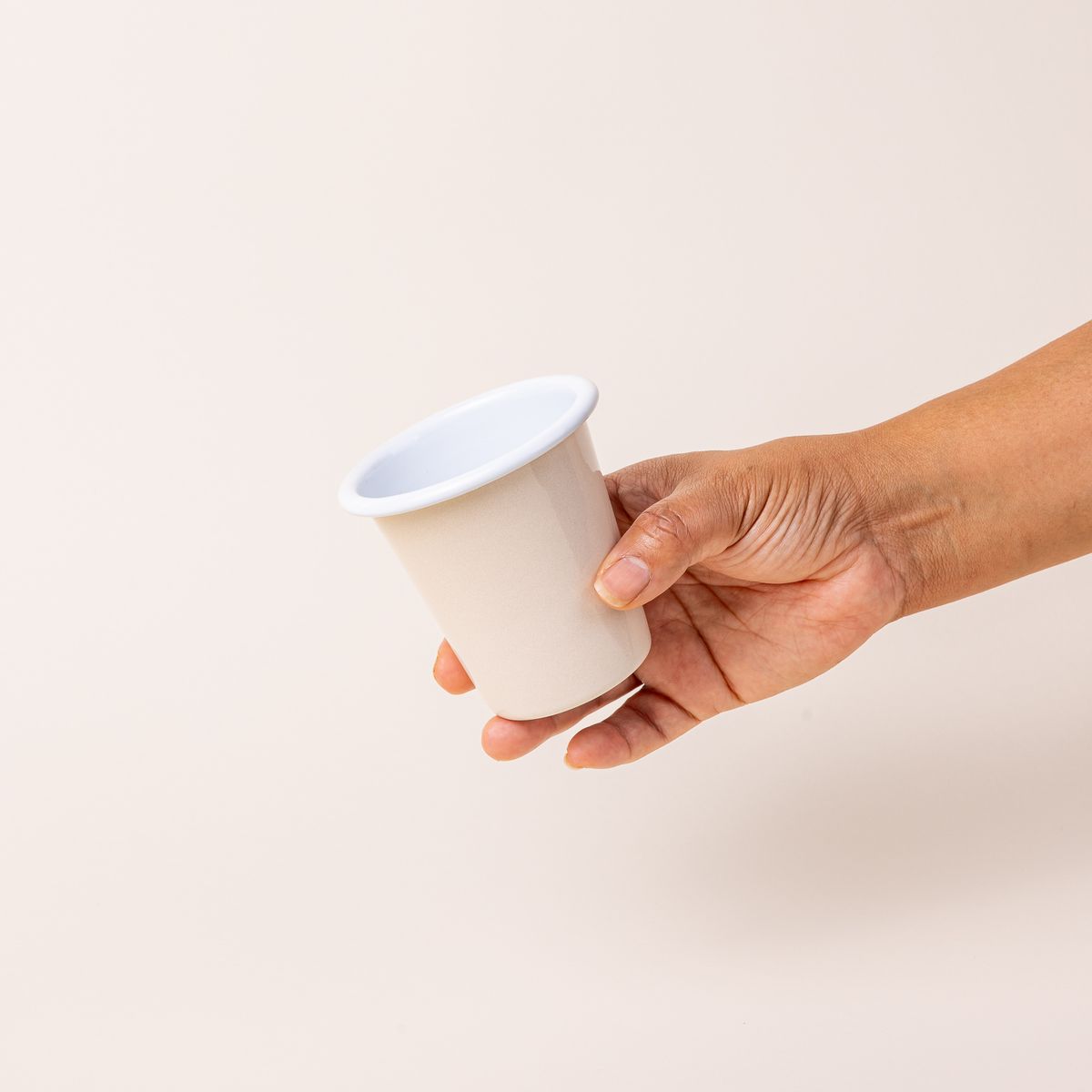 A hand holding a short enamel cup with a cream exterior and white interior