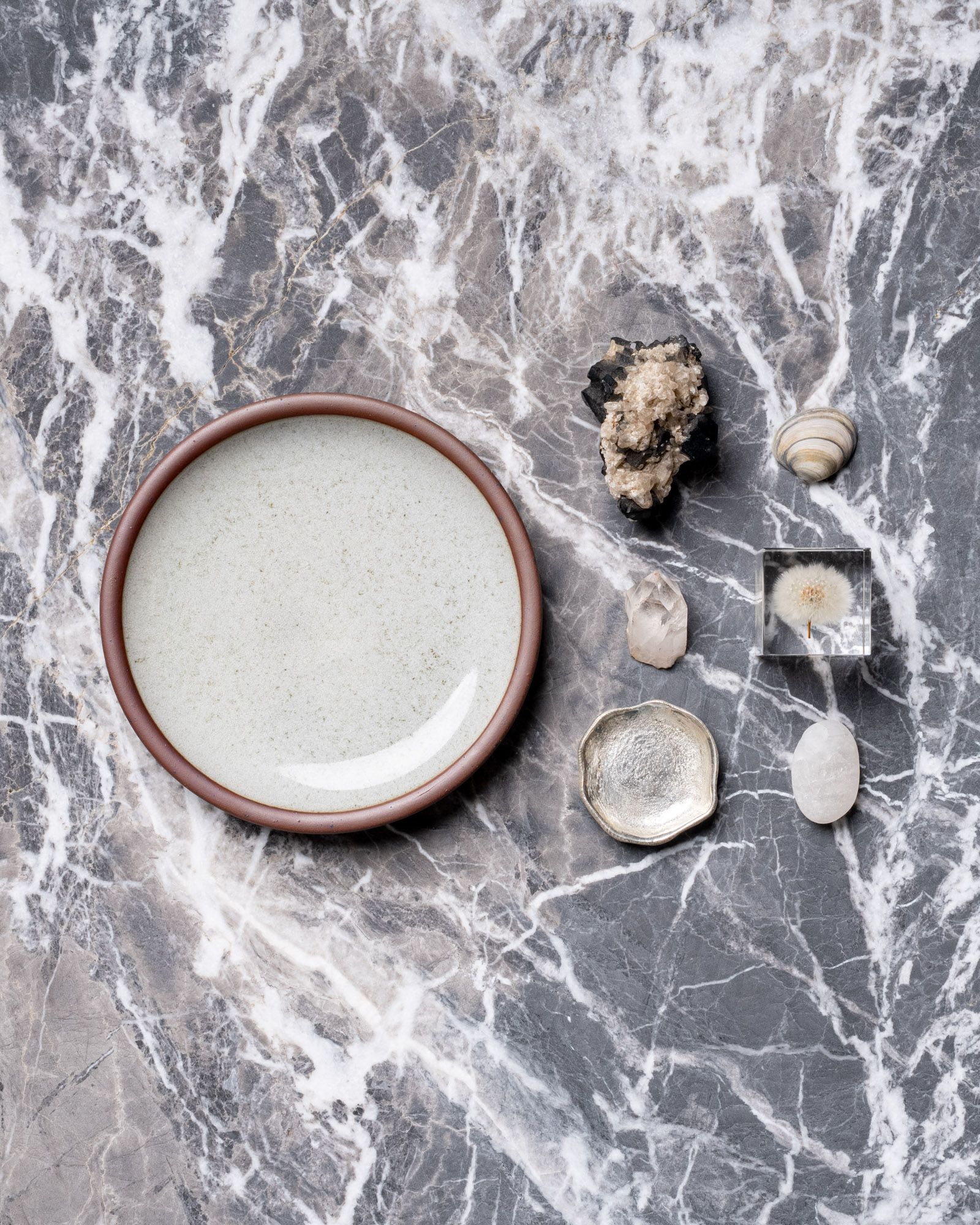 Round reflective icy white ceramic plate with unglazed rim on gray marble surface, arranged beside rocks, a shell, and small decorative objects.