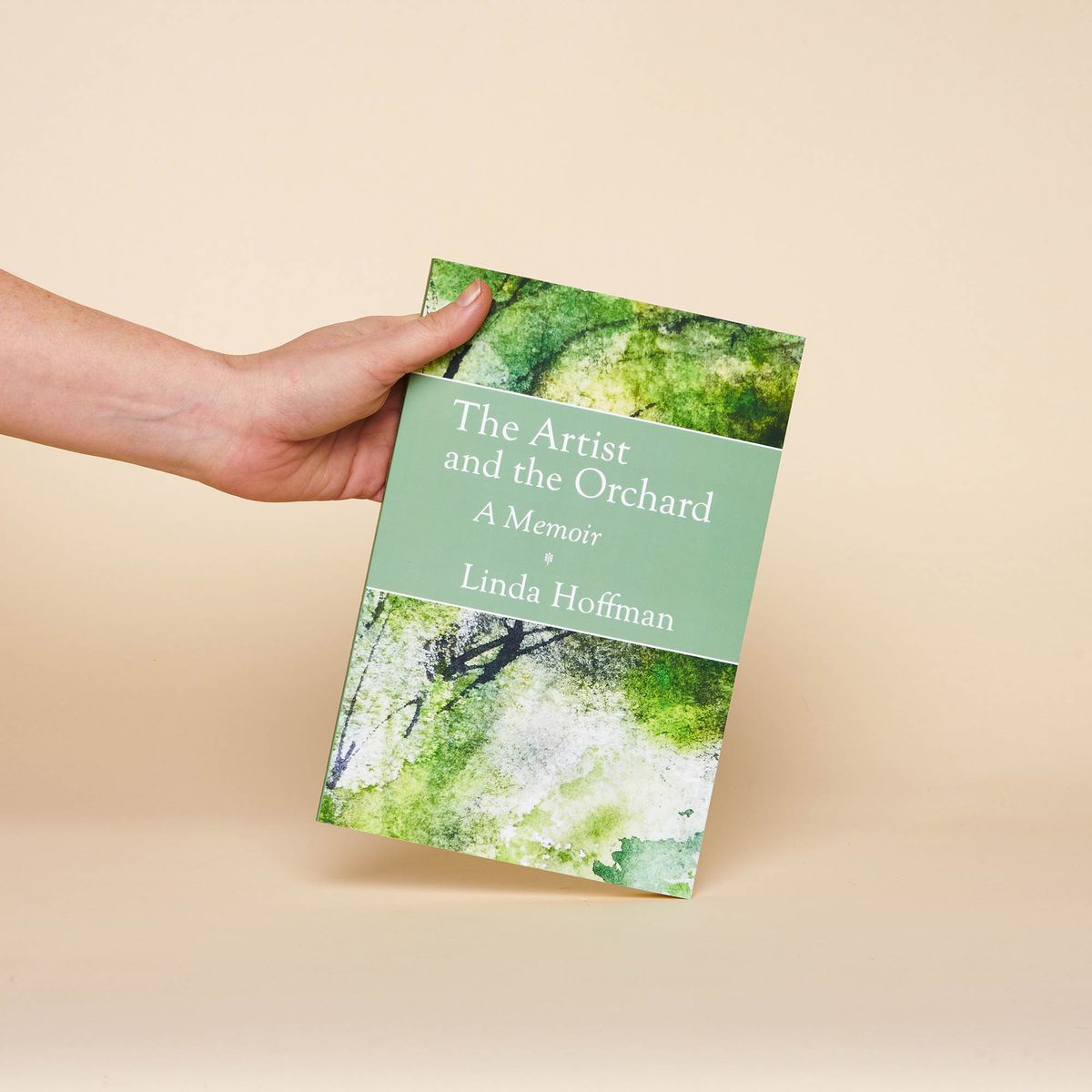 A hand holding The Artist and the Orchard: A Memoir by Linda Hoffman. Features an impressionist painting of trees.