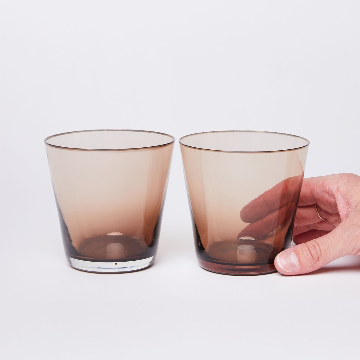Two glass tumblers that with a brown gradient that lightens towards the wider rim, with a hand holding the one of the right