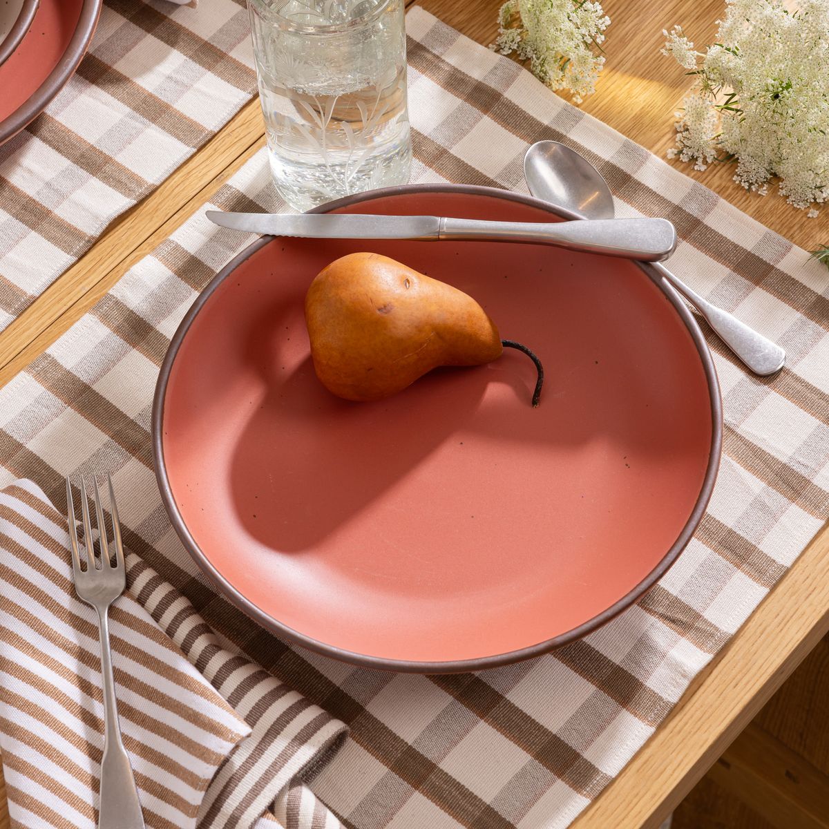 Watermelon pink ceramic plate with curved edge with a single brown pear, set on a checkered placemat with silverware, a glass of water, and flowers.