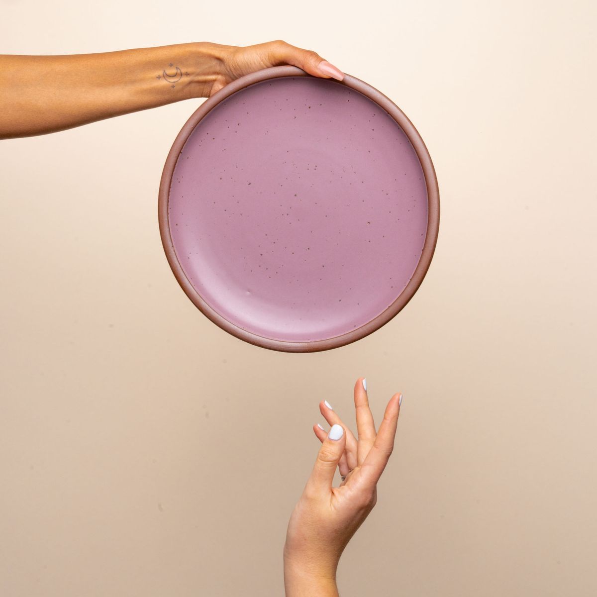 A hand holds a dinner sized ceramic plate in a soft mauve purple color featuring iron speckles and an unglazed rim. Another hand reaches for it.