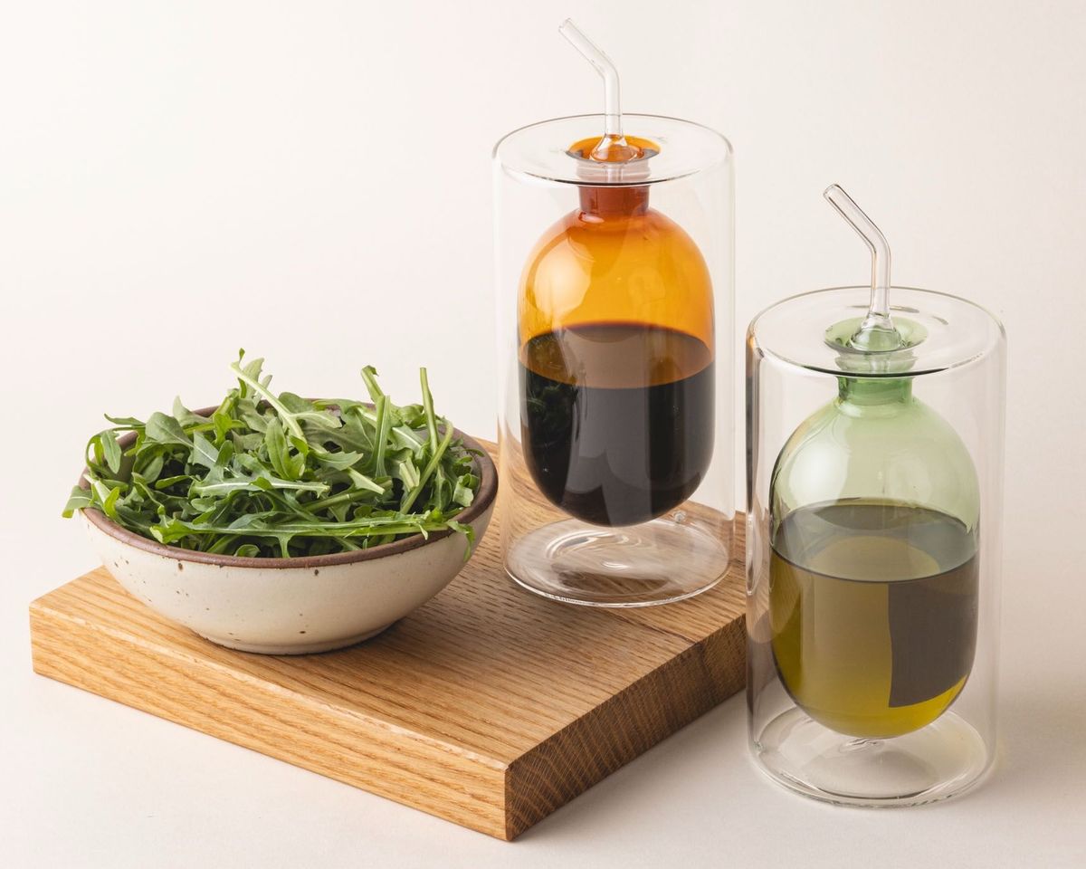 Two artful glass cruets in a an orange and light green color. There is a rounded bulb that acts as a container for the cruet, surrounded by a doubled glass wall. The cruets accompany a small salad bowl and wooden cutting board