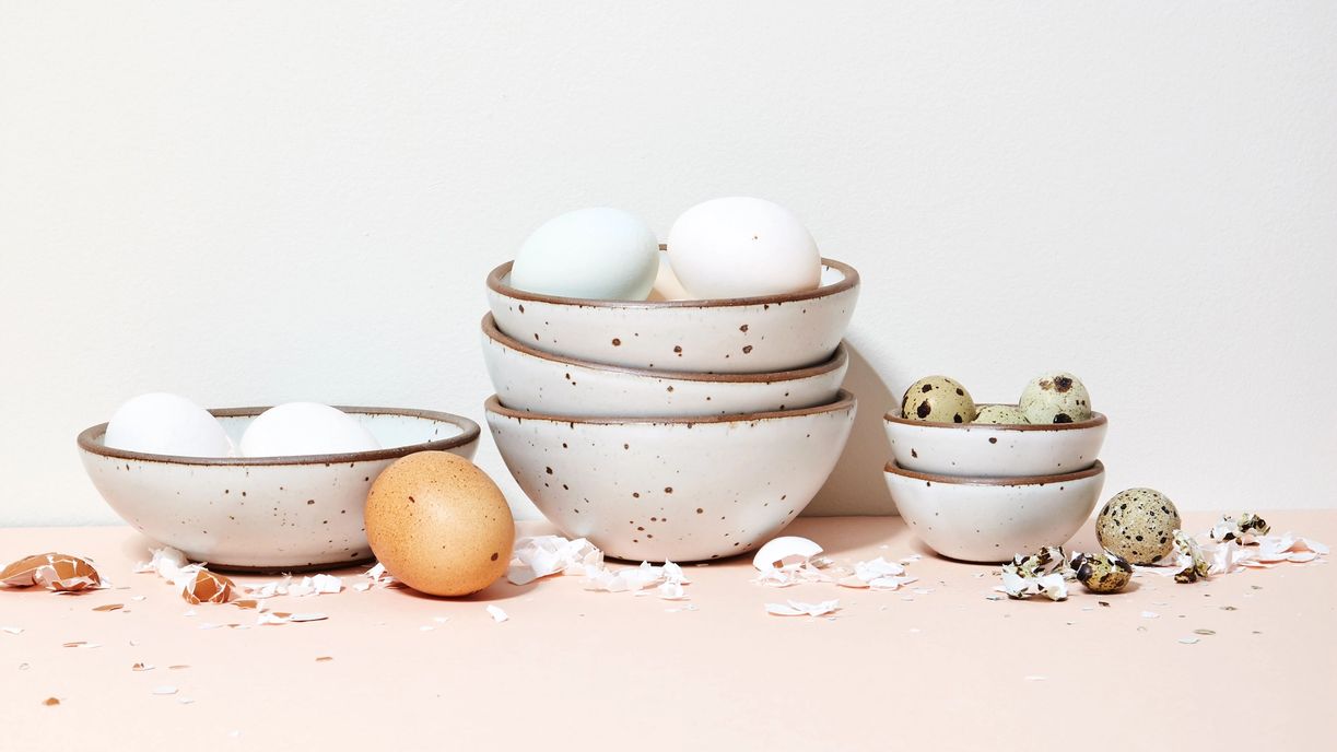 Stacks of various sizes of ceramic bowls in a cool white color. Different kinds of eggs fill each bowl with crushed shells artfully arranged around.