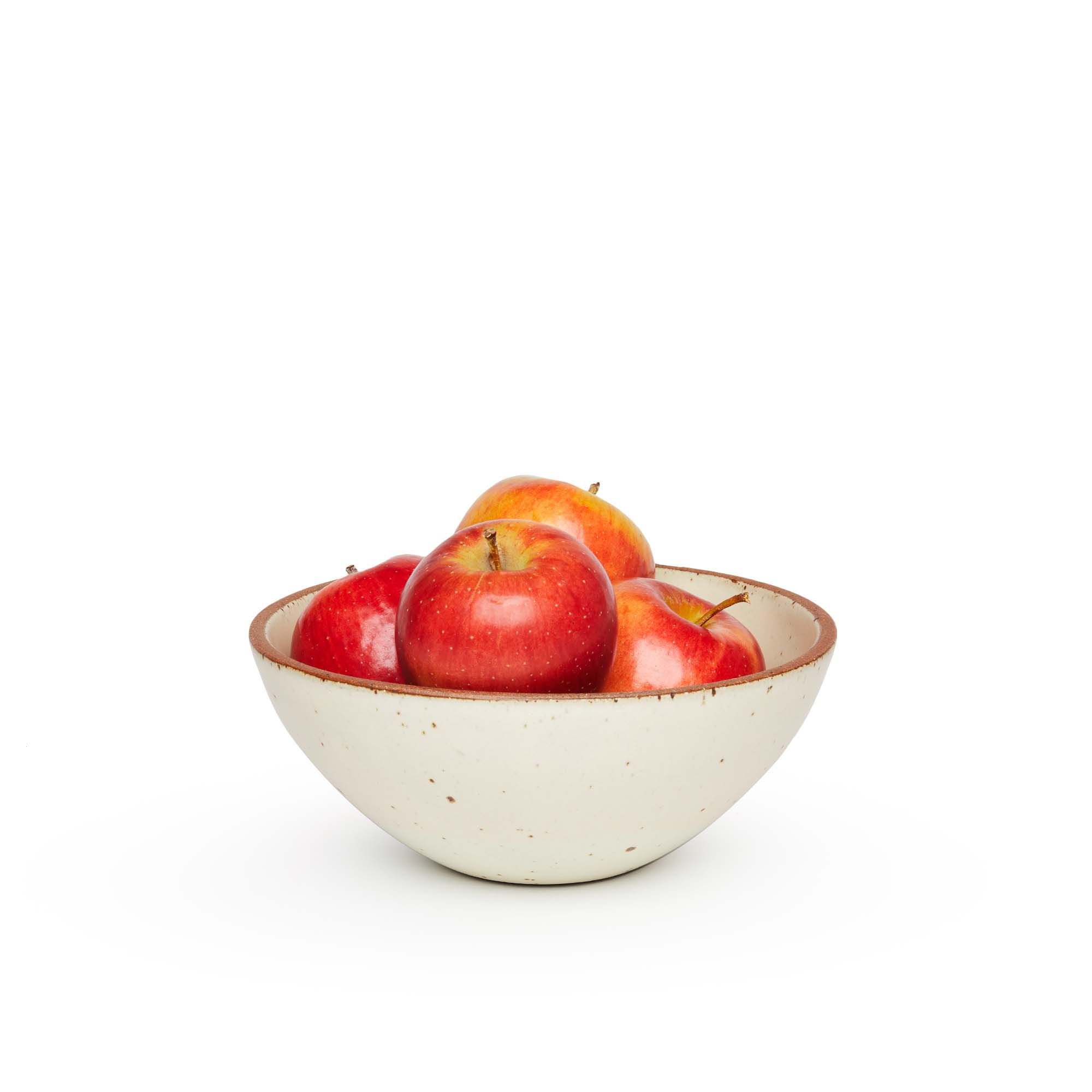 A medium rounded ceramic bowl in a warm, tan-toned, off-white color featuring iron speckles and an unglazed rim, filled with apples