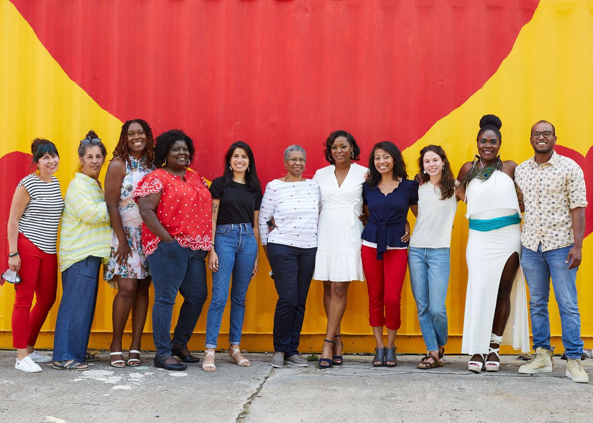 A diverse group of several people line up and smile at the camera in front of mural with yellow and red colors