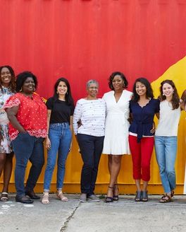A diverse group of several people line up and smile at the camera in front of mural with yellow and red colors