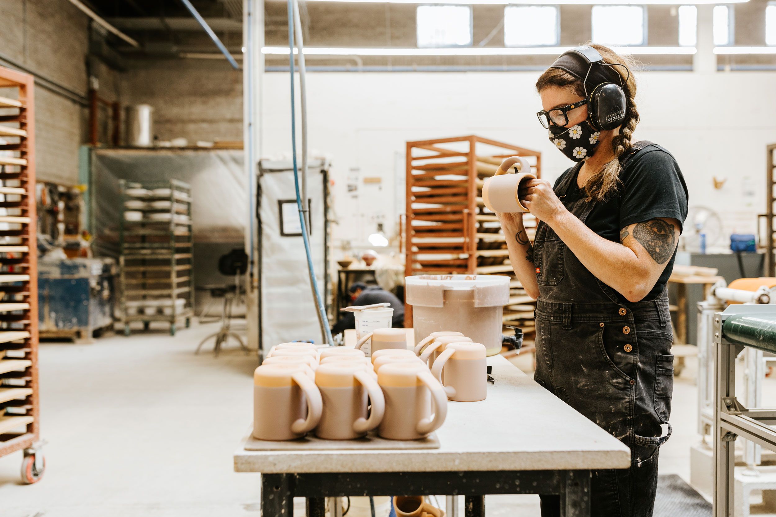 A person wearing safety headphones and a mask is working on mug and standing in front of a table of unglazed mugs.