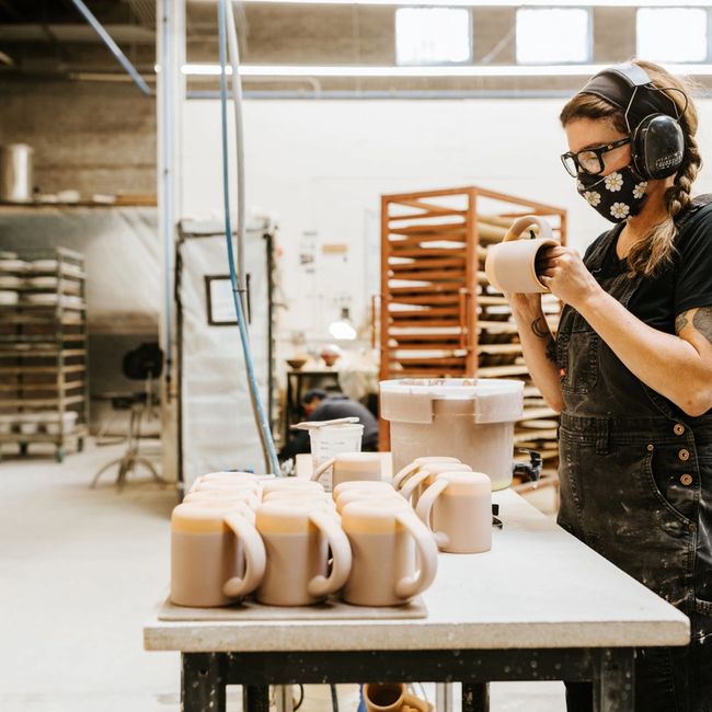 In a warehouse setting, a worker is holding an East Fork mug in progress, with a table of mugs in front of them filled with unglazed mugs.