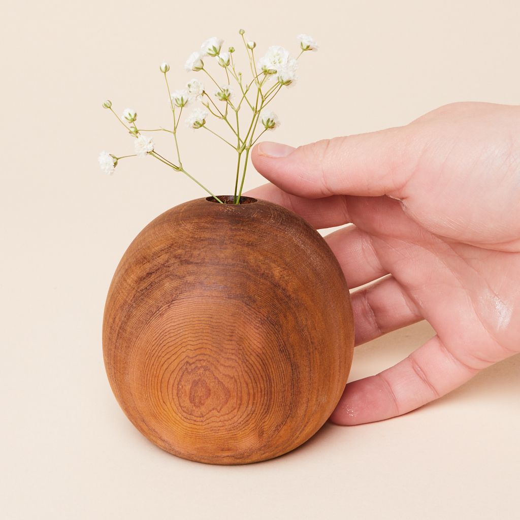 Three sprigs of tiny white flowers on thin green stems stick out of the hole at the top of an orb-shaped brown vase made of wood