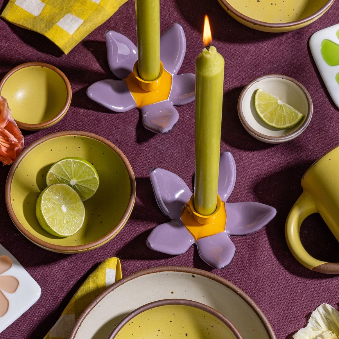A sculptural glass purple flower candleholder with an orange center with a neon green taper candle, and on a colorful yellow and purple table.