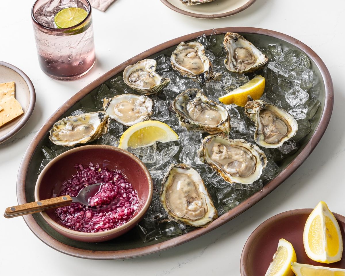 A large forest green oval ceramic platter filled with oysters on ice, and a plum bowl with sauce and a spoon