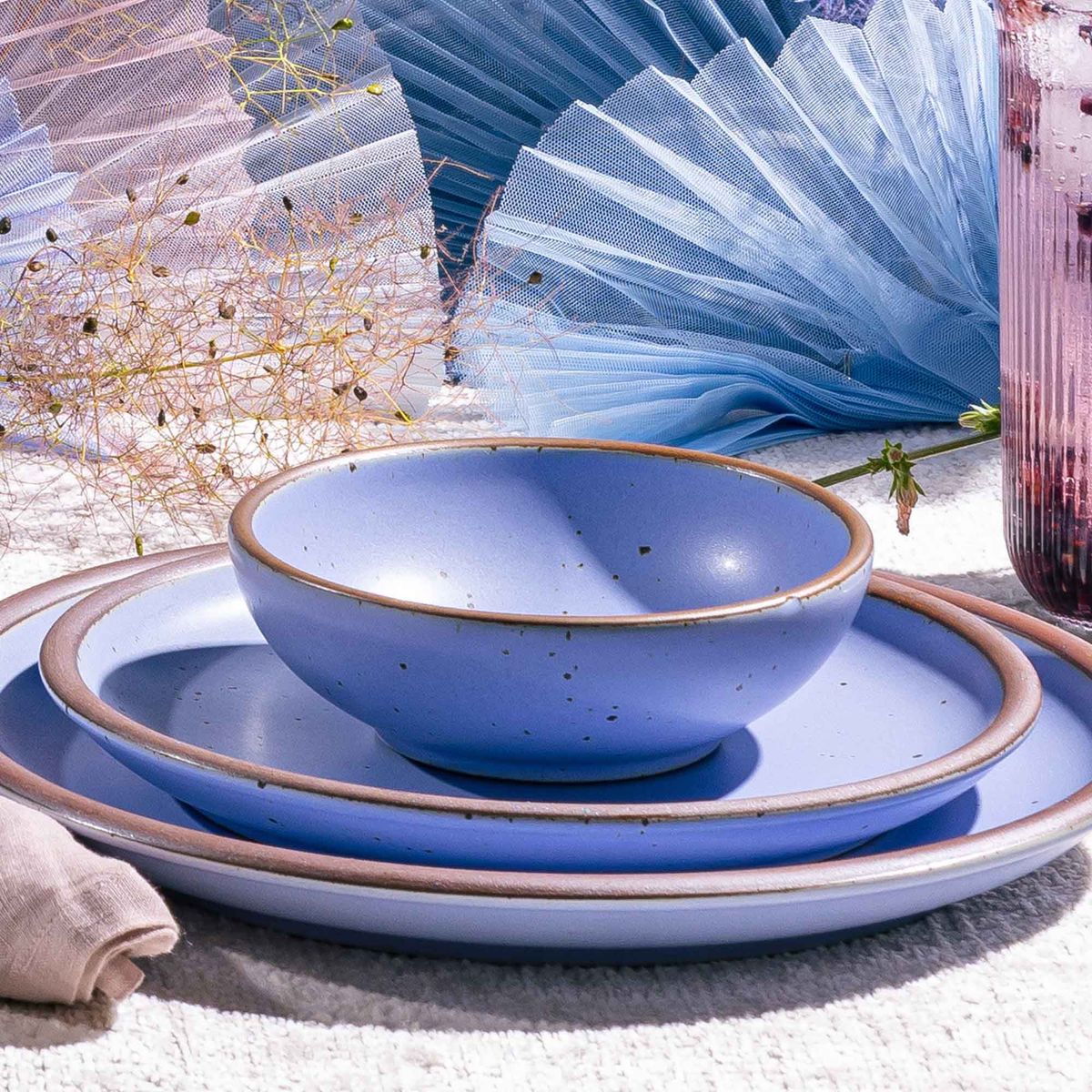 A place setting with periwinkle dinner plates and a shallow bowl and fabric sculpture in the background