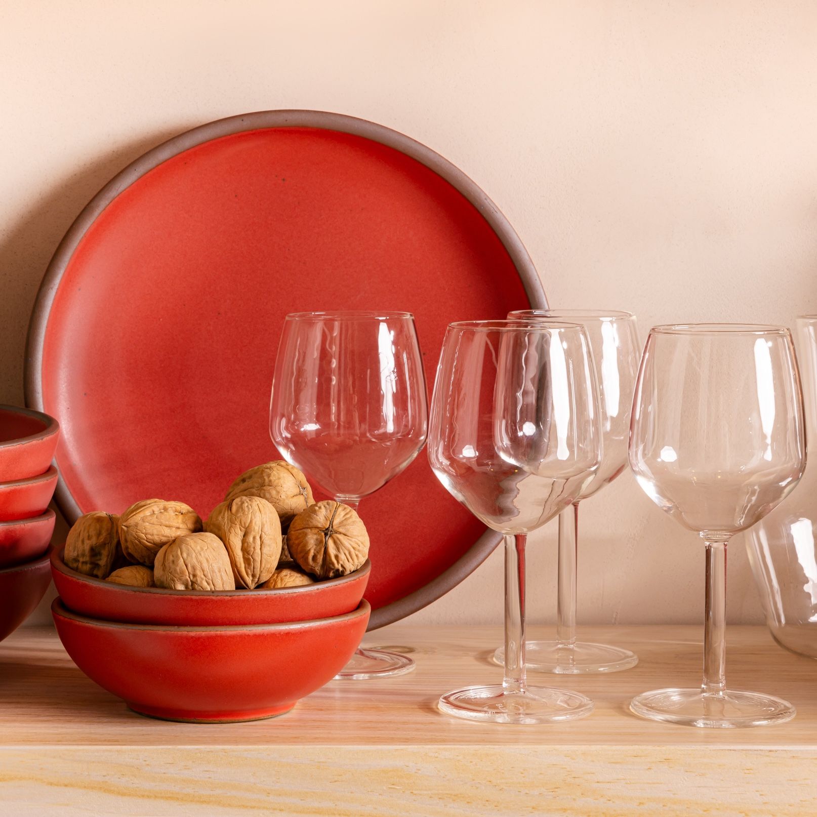 On a shelf is several clear simple wine glasses, a propped up red plate, and a stack of small red bowls with walnuts inside