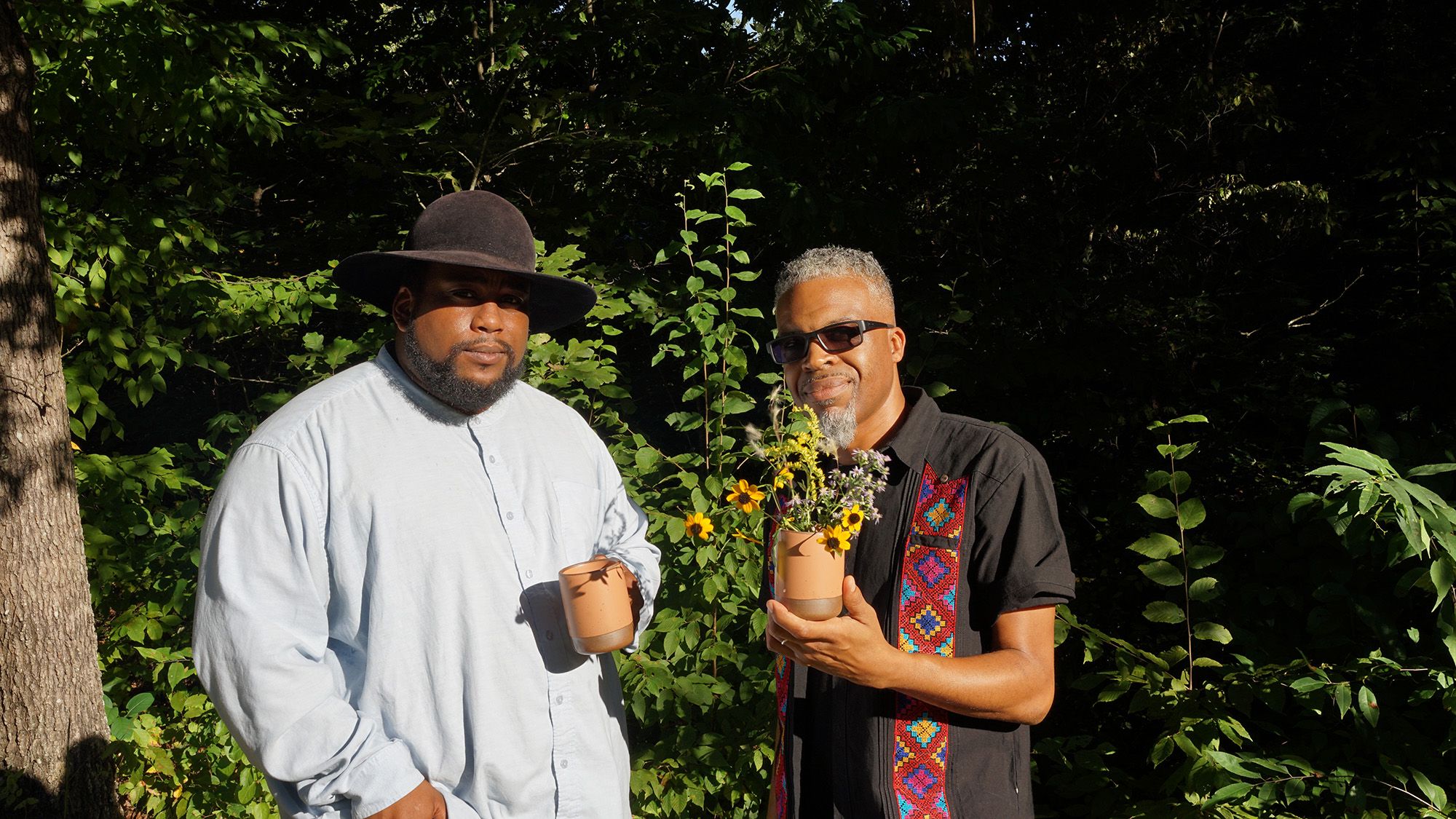 Two people standing in bright sun in front of green foliage, one wearing a light blue shirt with a black hat holding a mug and another wearing a black shirt and sunglasses holding a mug full of wildflowers