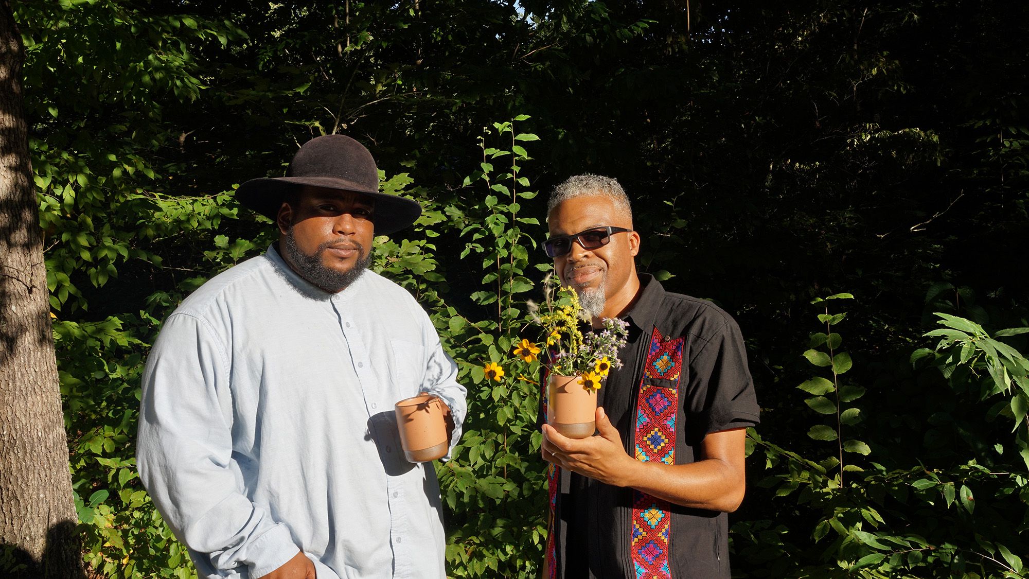 Two people are outside staring at the camera - the left person is wearing a button down shirt and hat and holding a mug, the other is wearing sunglasses and button down shirt, holding a mug that holds wildflowers