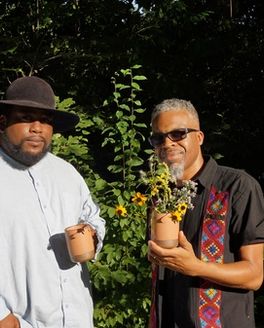 Two people are outside staring at the camera - the left person is wearing a button down shirt and hat and holding a mug, the other is wearing sunglasses and button down shirt, holding a mug that holds wildflowers