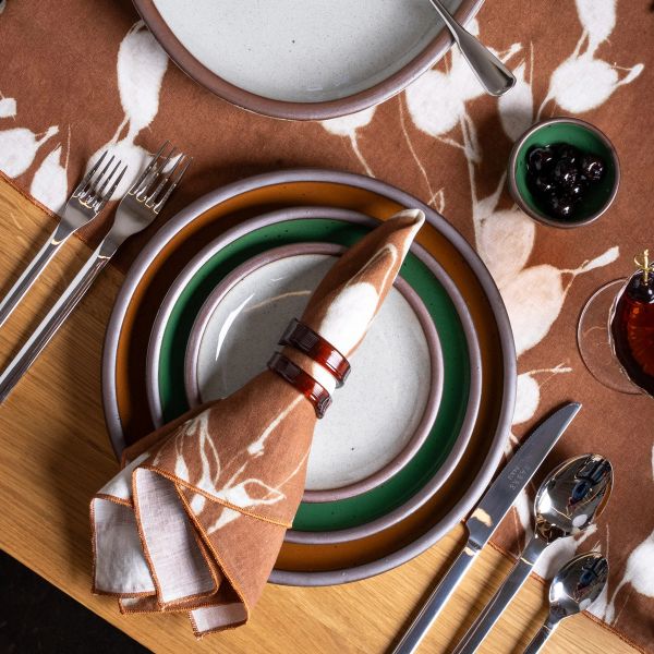 Table setting with layered ceramic plates in a reflective icy white, glossy green, and glossy brown on a brown floral table runner, with silverware and a folded napkin.
