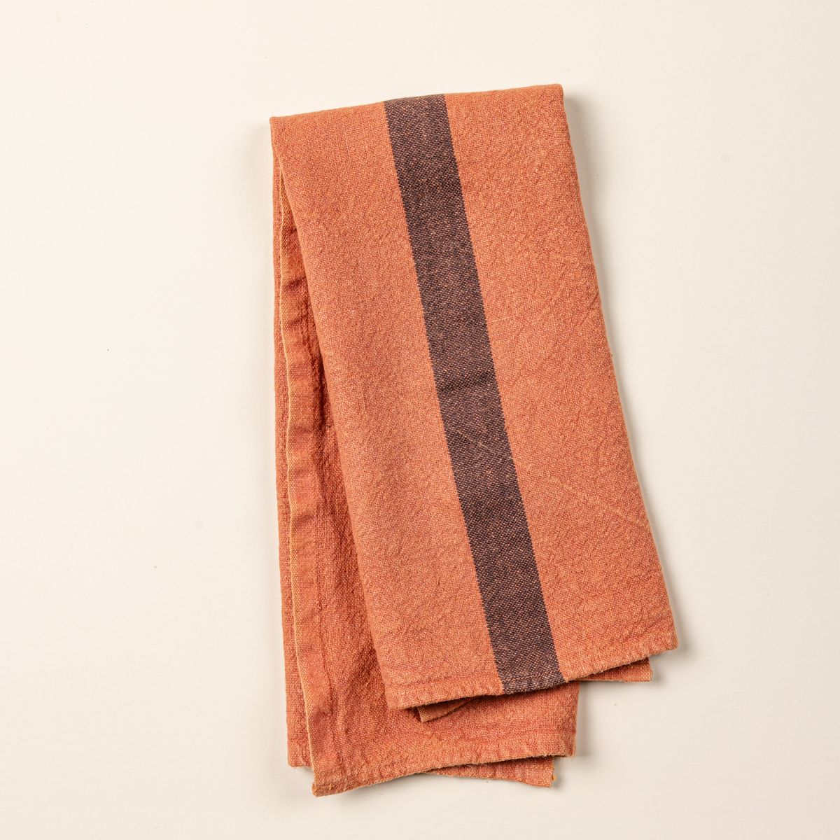 A folded linen towel in a terracotta color with a plum stripe in the middle