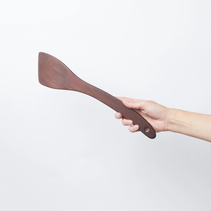 Hand holding a black walnut wood spatula with a slanted head and a hole at the bottom 