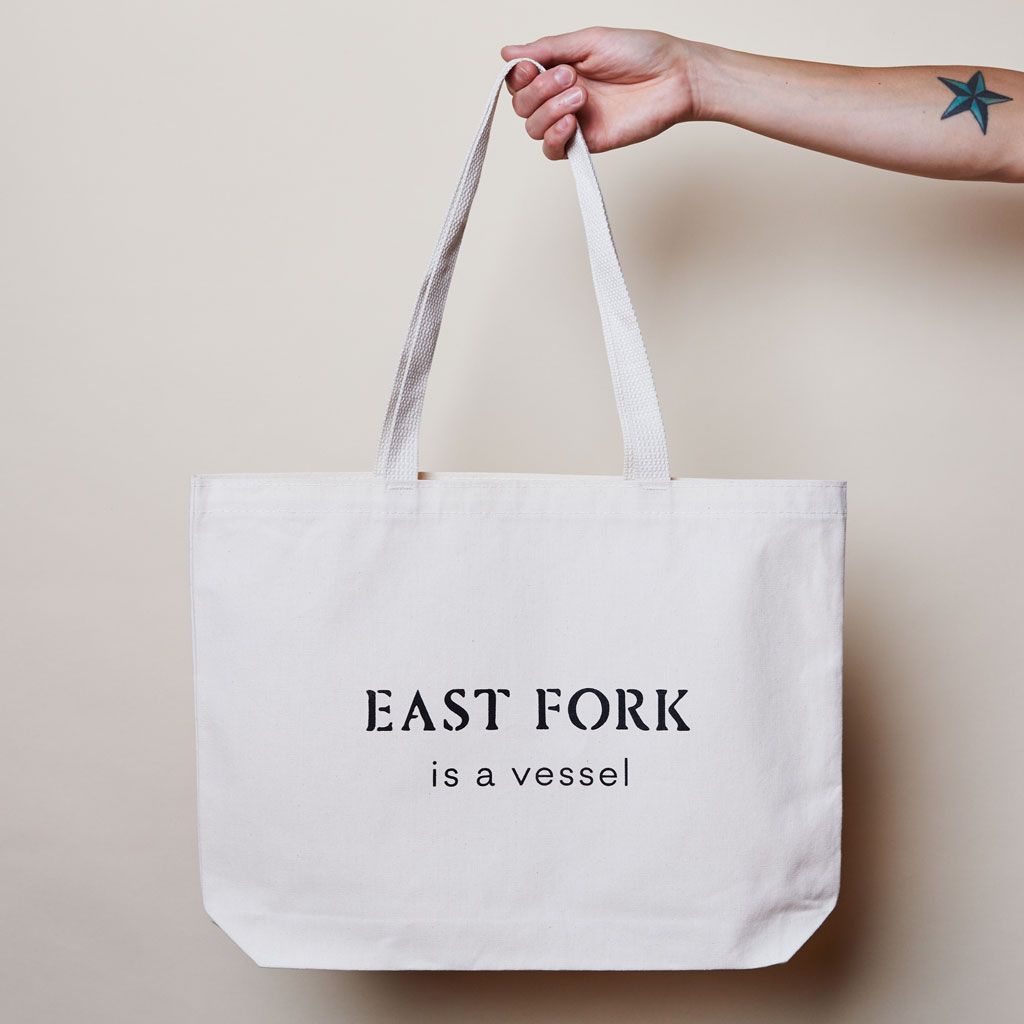 Hand holds the handle of an East Fork tote bag, which reads "East Fork is a vessel"