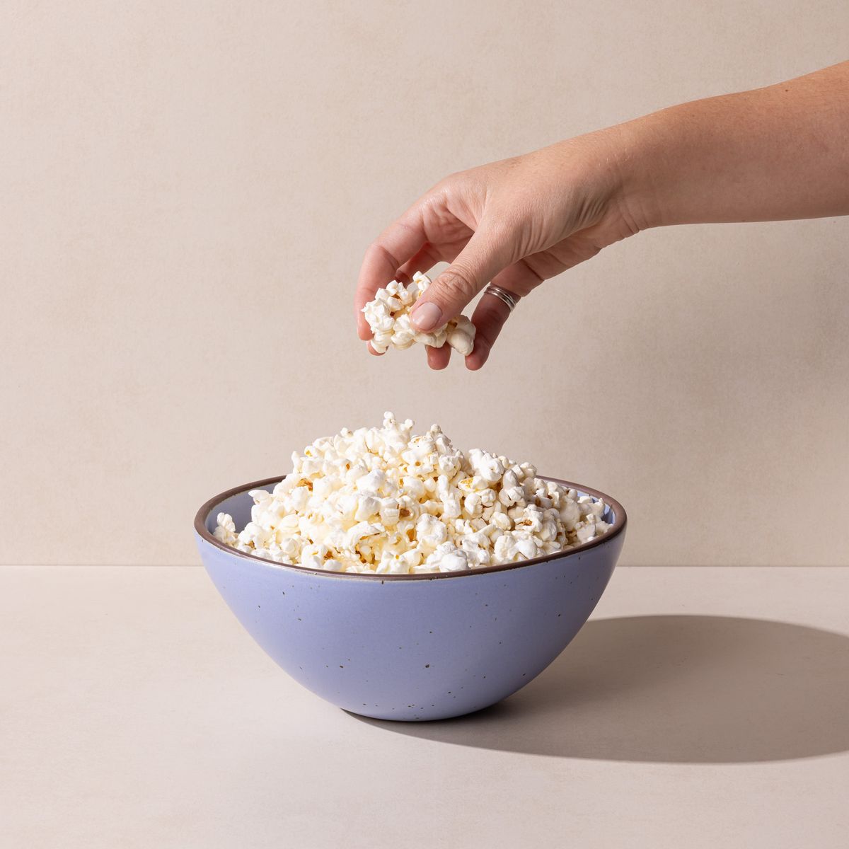 A hand is reaching for popcorn that fills a large rounded ceramic bowl in a periwinkle color featuring iron speckles and an unglazed rim