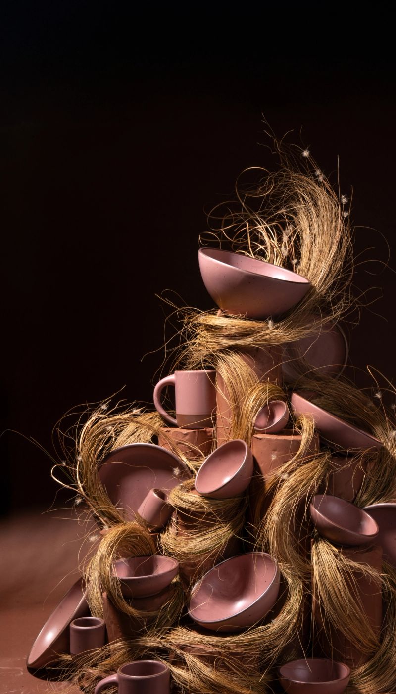 Stack of dusty mauve pink ceramic bowls, plates, and mugs piled together with strands of dried grass against a dark background.