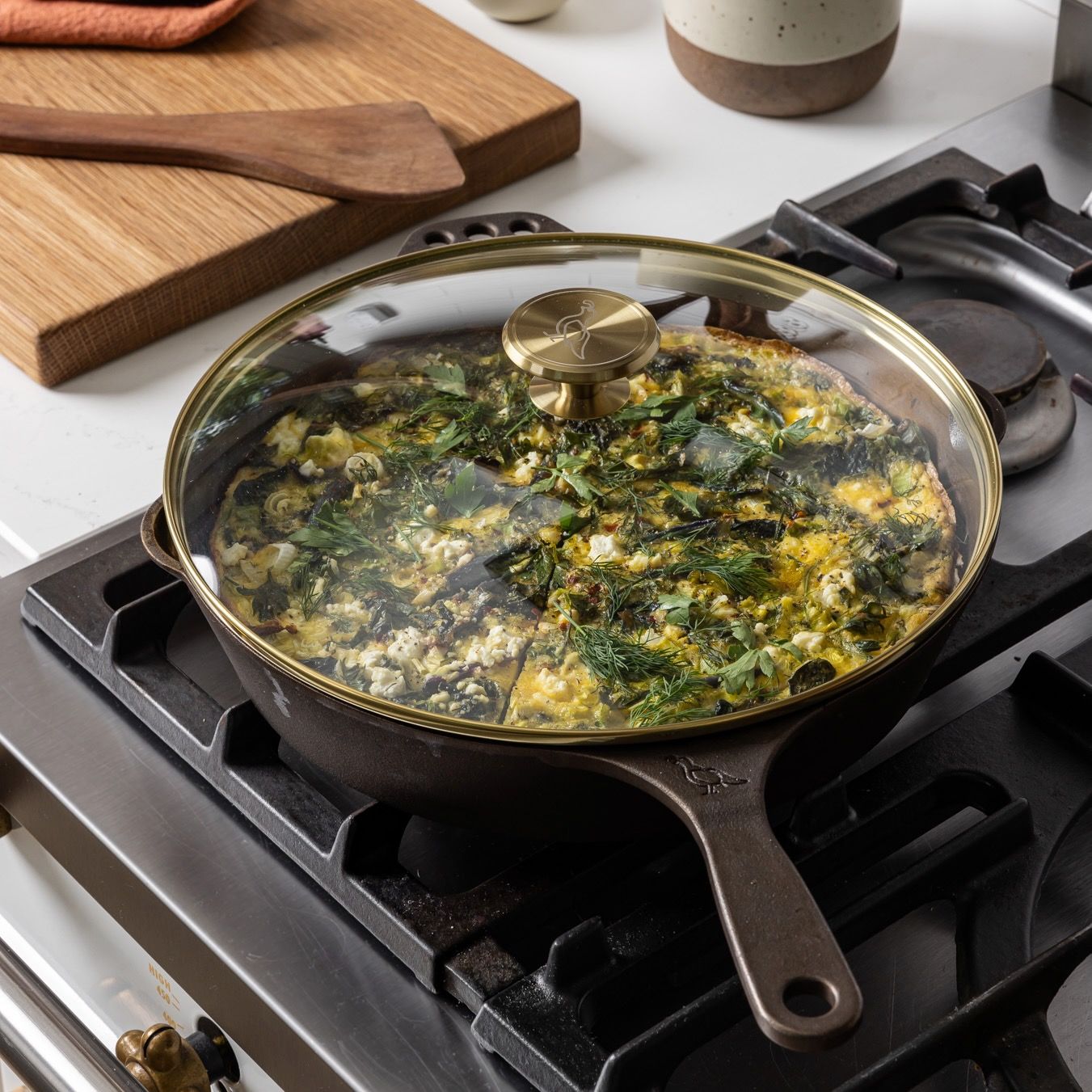 On a stove, there's  glass skillet lid with a brass lip and brass handle knob over a cast iron skillet. The skillet is filled with a casserole.