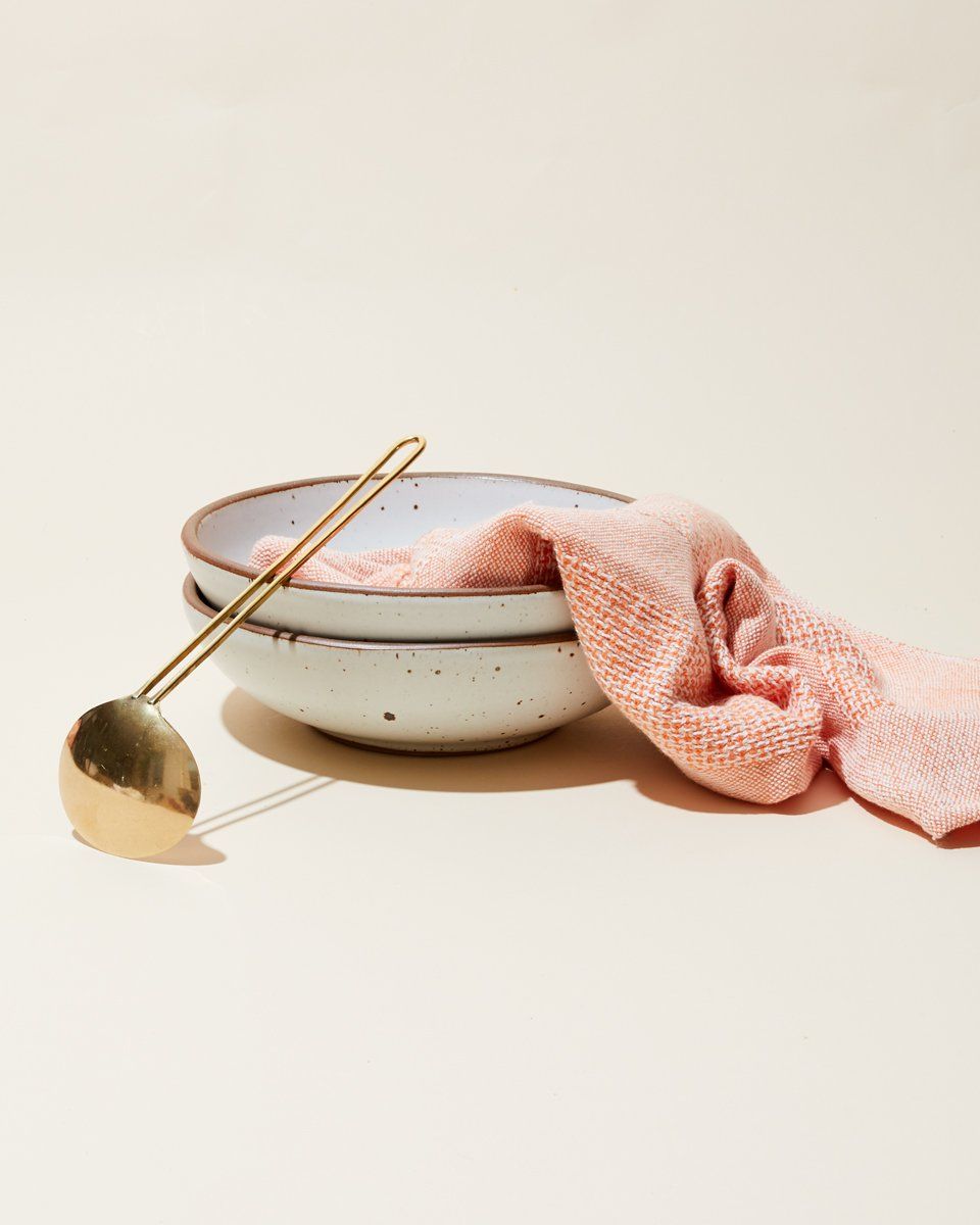2 shallow dinner bowls in a white color are stacked and are with a woven salmon hand towel and a brass soup spoon