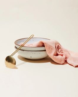 2 shallow dinner bowls in a white color are stacked and are with a woven salmon hand towel and a brass soup spoon