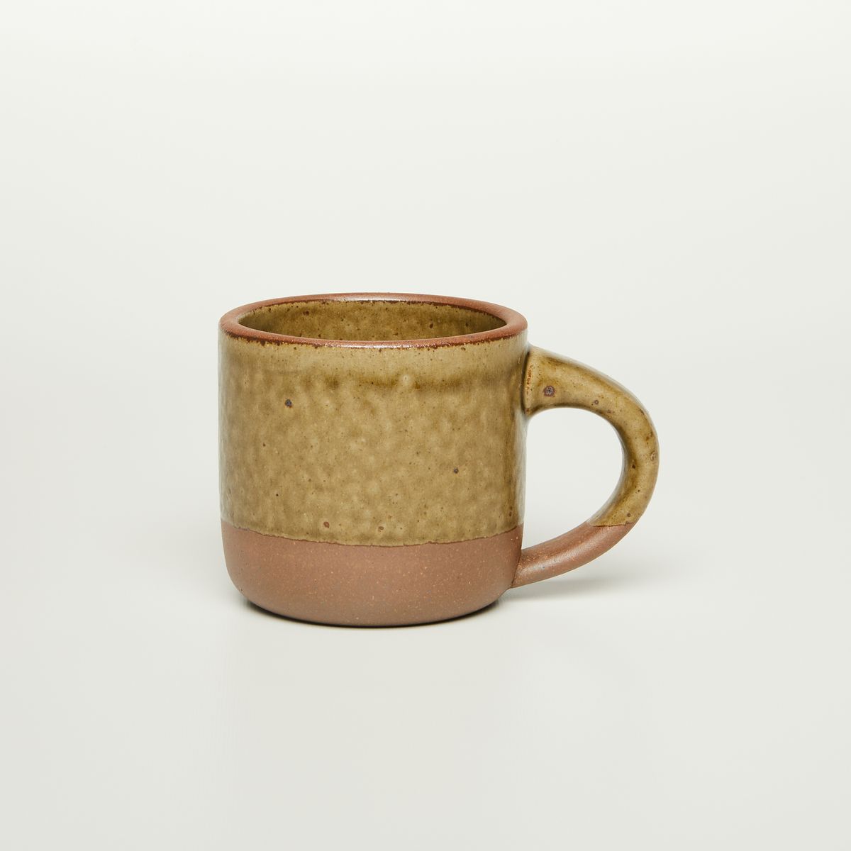 A small sized ceramic mug with handle in an earthy green and brown color featuring iron speckles and unglazed rim and bottom base.