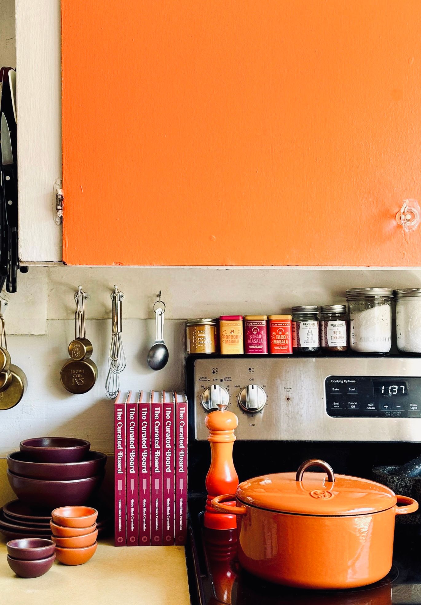 A stovetop and counterspace filled with kitchen tools and colorful ceramic bowls in a bold orange and plum, as well as an orange dutch oven.