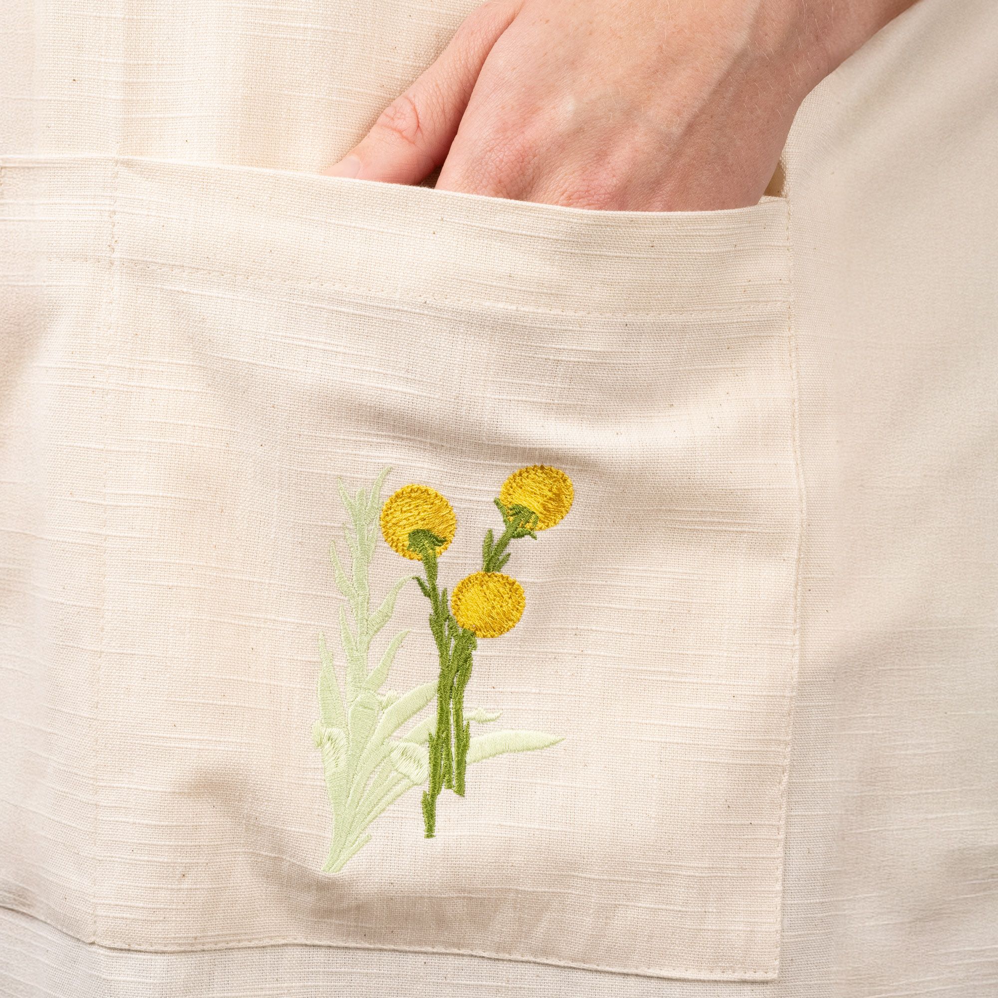 A hand in a canvas apron pocket with a yellow embroidered flower.