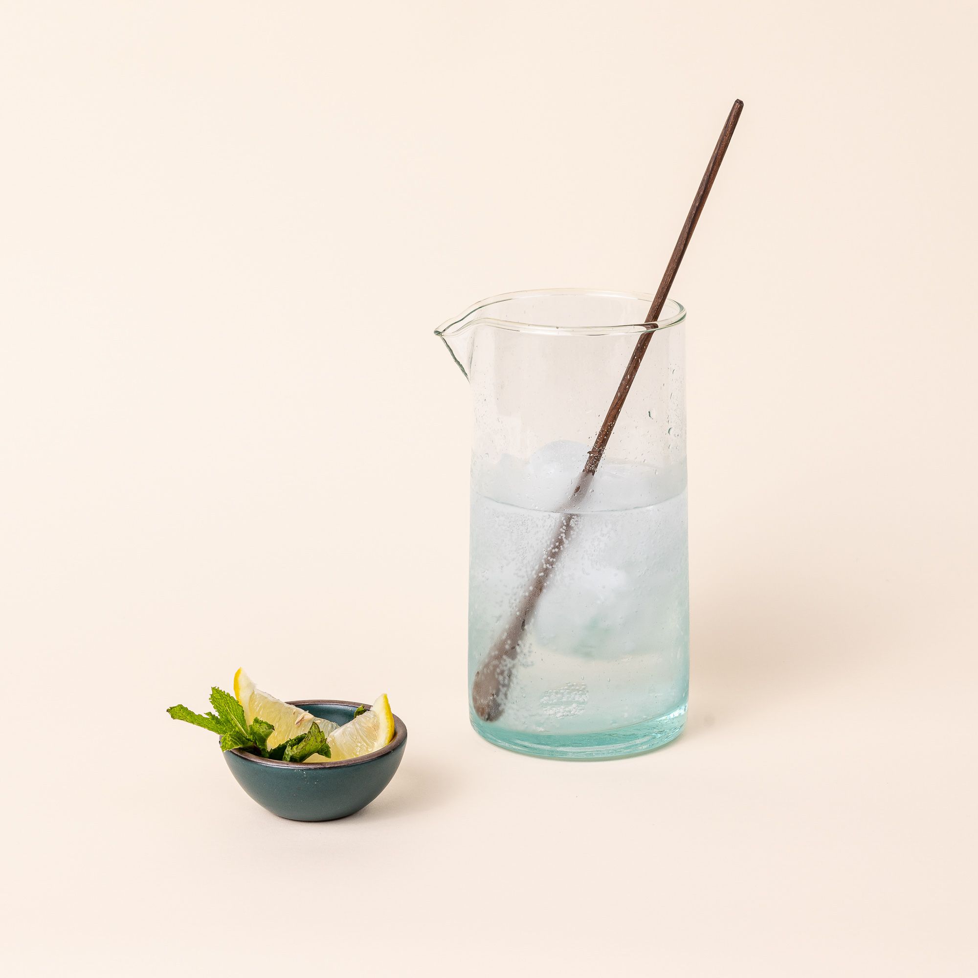 A long thin walnut cocktail stirrer with a small spoon-like shape on the end sits inside a wide clear pitcher filled with a clear liquid and ice. A tiny teal bowl sits next to it with sliced lemon and mint.