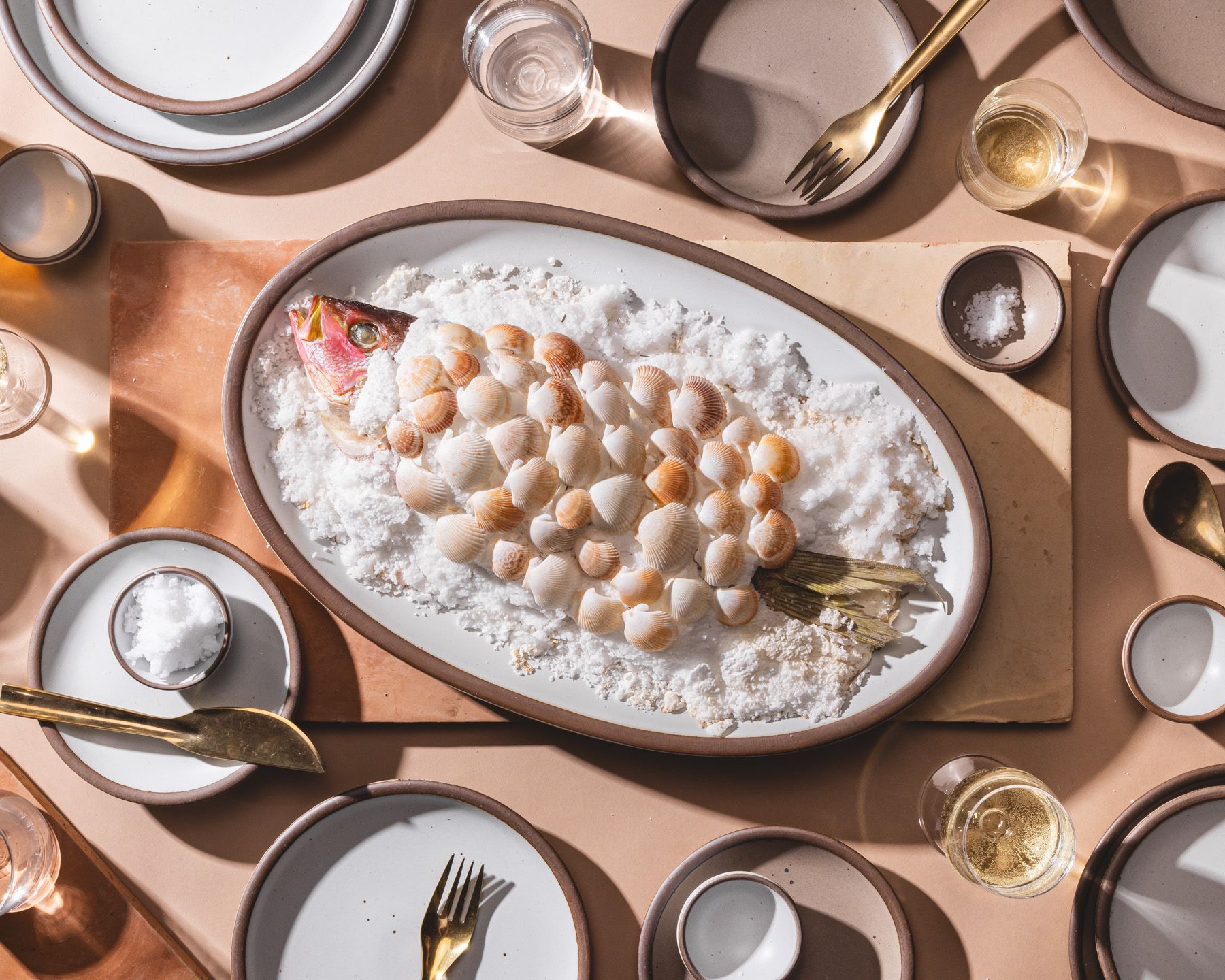 A large oval platter in white with a stunning fish dish on top surrounded by plates and brass flatware