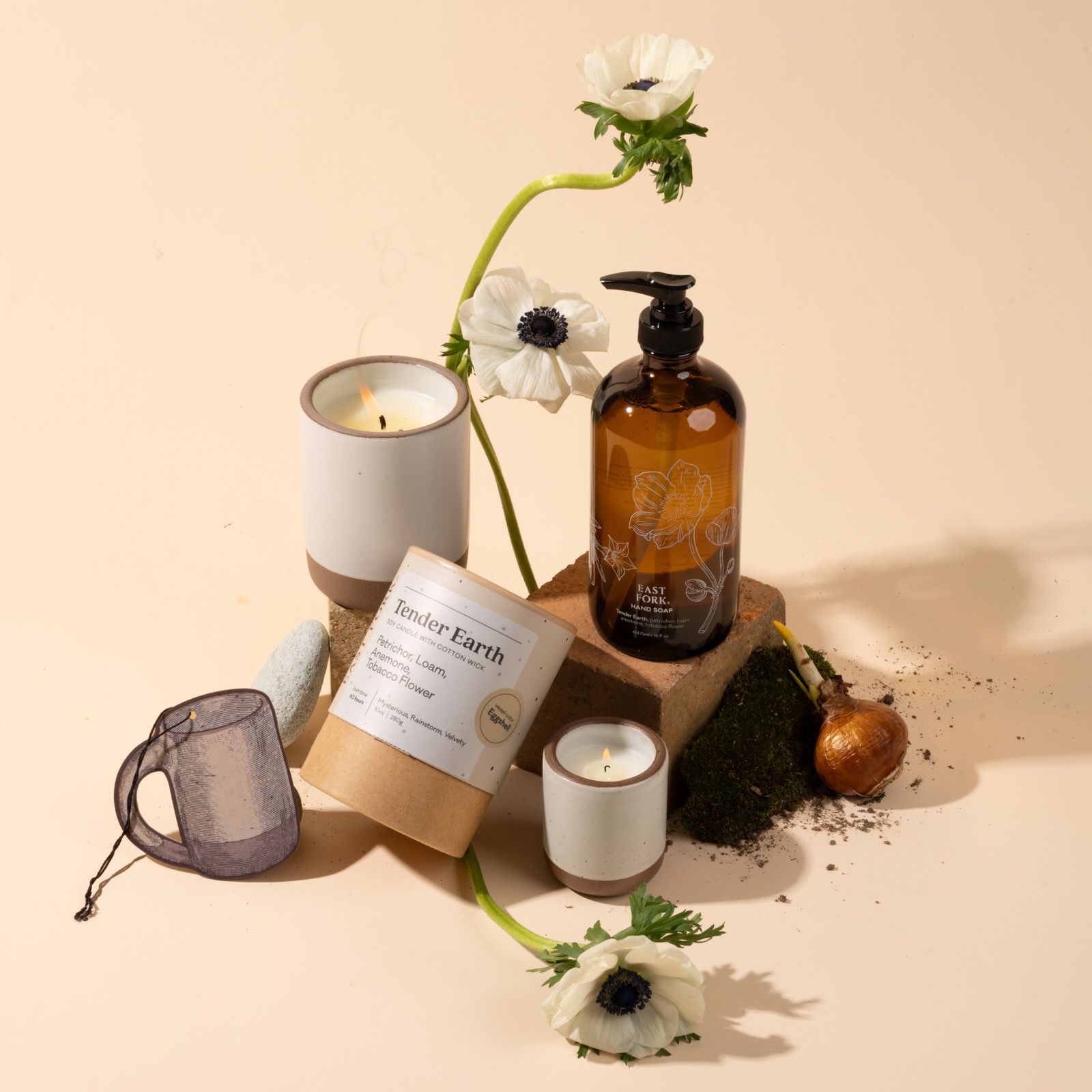 Hand soap bottle and lit candles arranged with white flowers, moss, soil, and a candle jar on a neutral background.