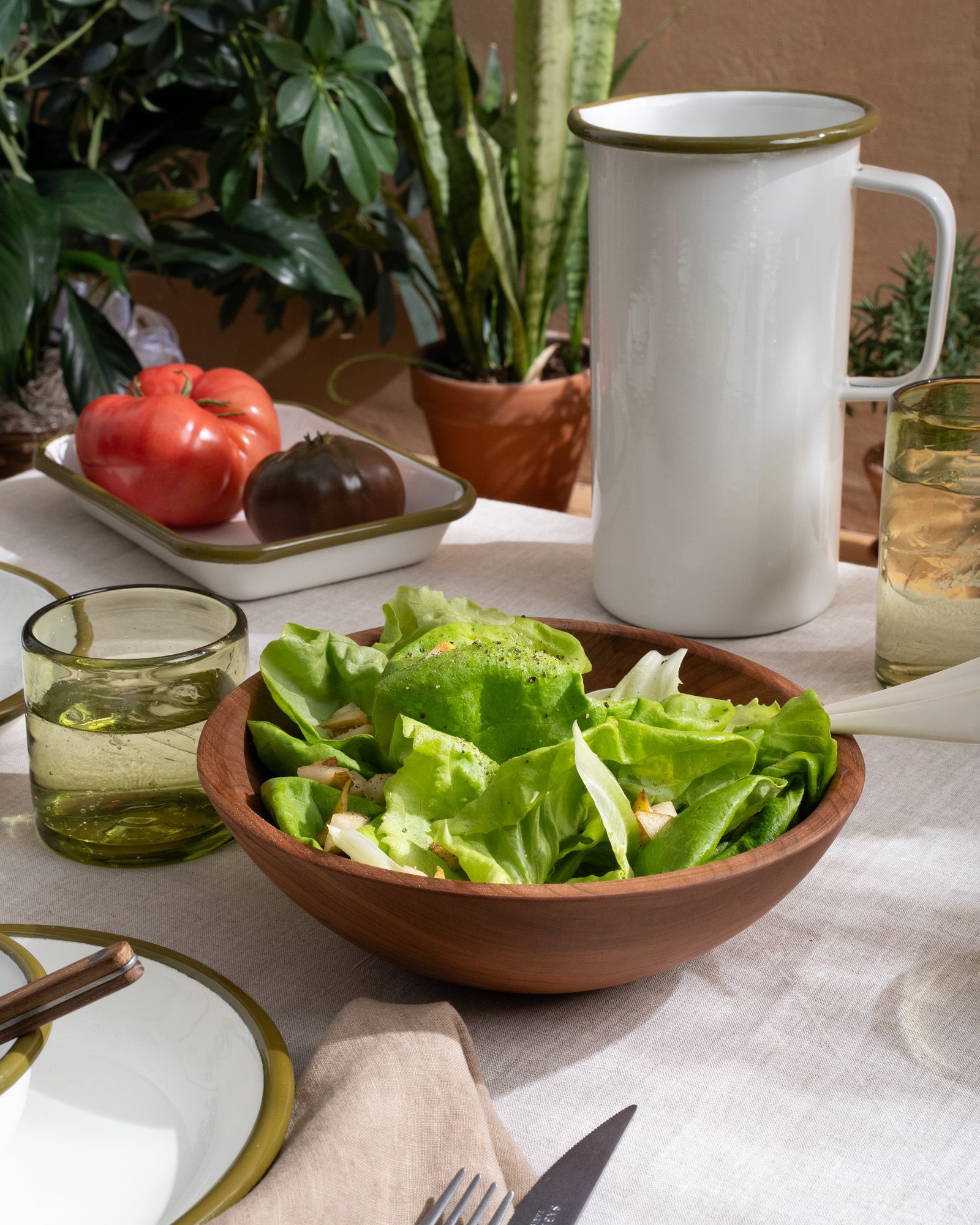 A turned walnut wood bowl filled with salad on a full table with an enamel pitcher and dinnerware, and green glasses.