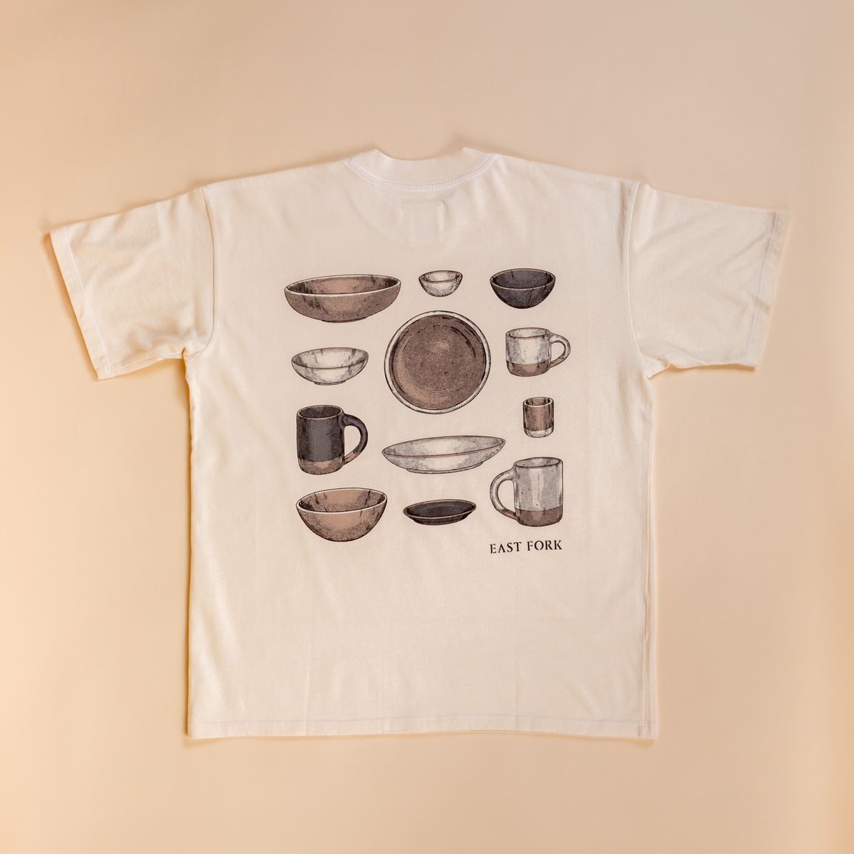 Flat lay of the back of a t-shirt in cream against a cream background. The back has an illustration of various mugs, plates, and bowls all in neutral colors.