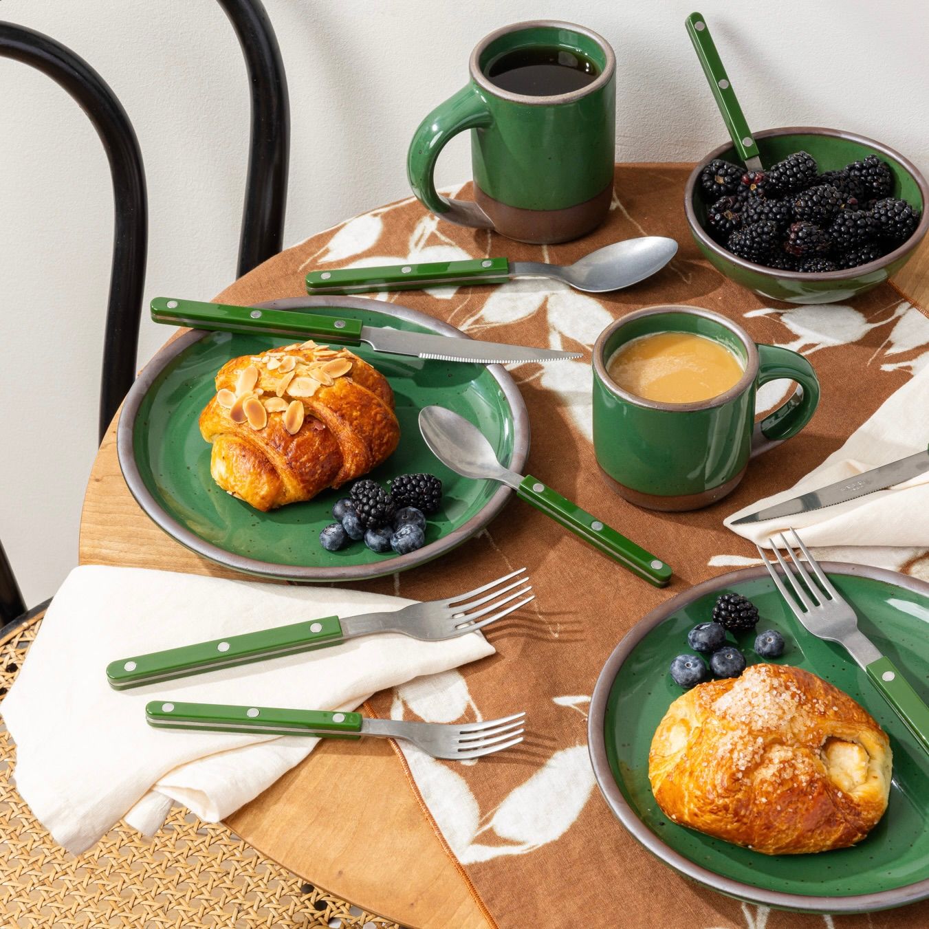 Breakfast table with glossy emerald green mugs and plates, croissants, berries, and coffee on a floral-patterned tablecloth.