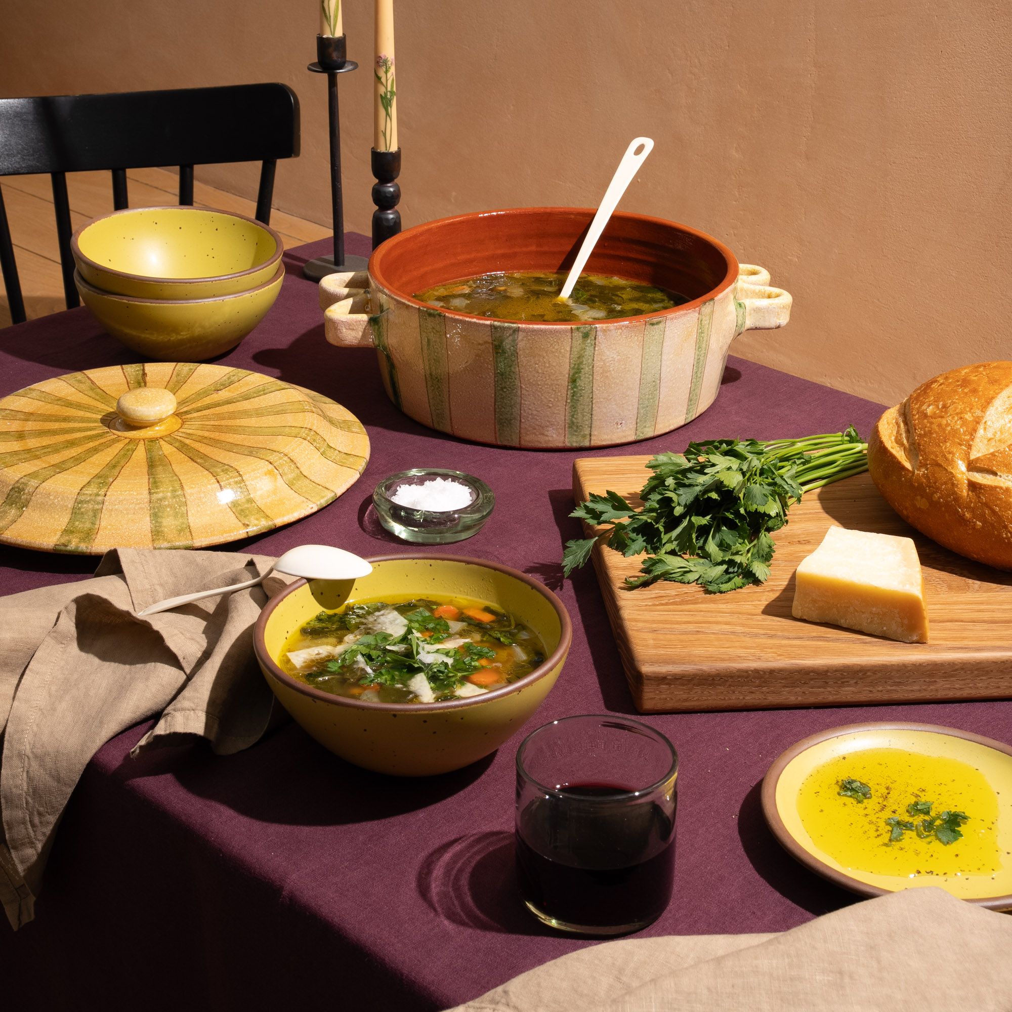A table with with a large striped terracotta casserole dish, yellow soup bowls, a cutting board with bread, and more.