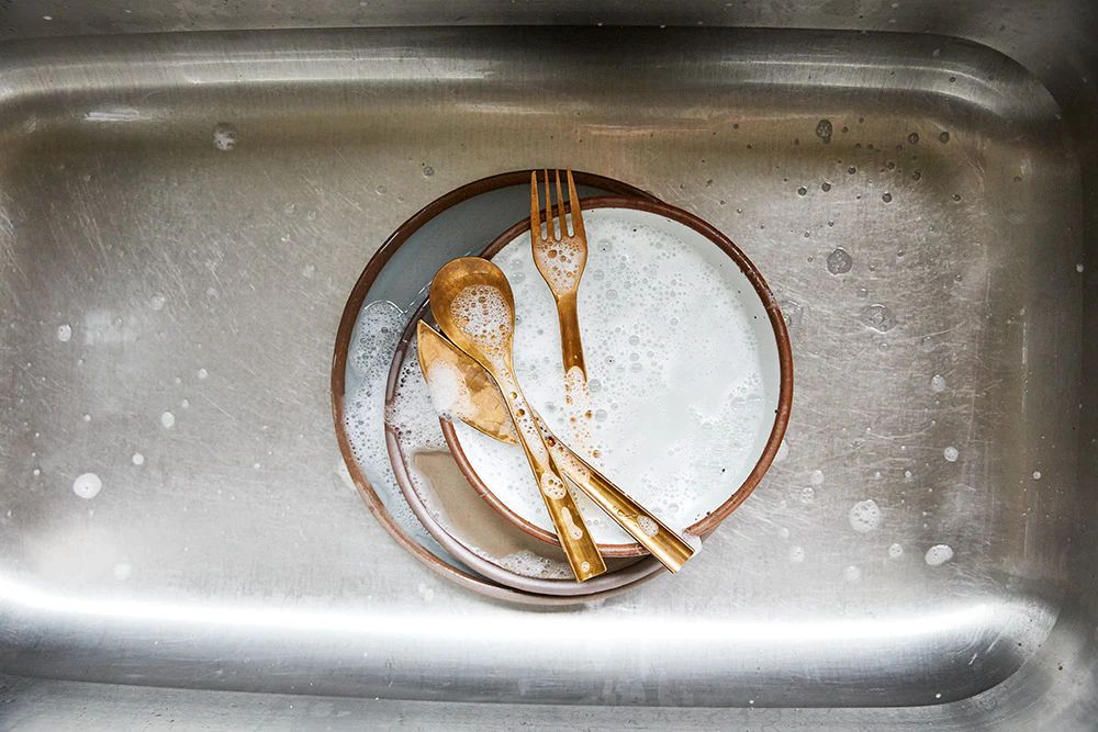 Brass cutlery in a sink on top of sudsy East Fork dishes