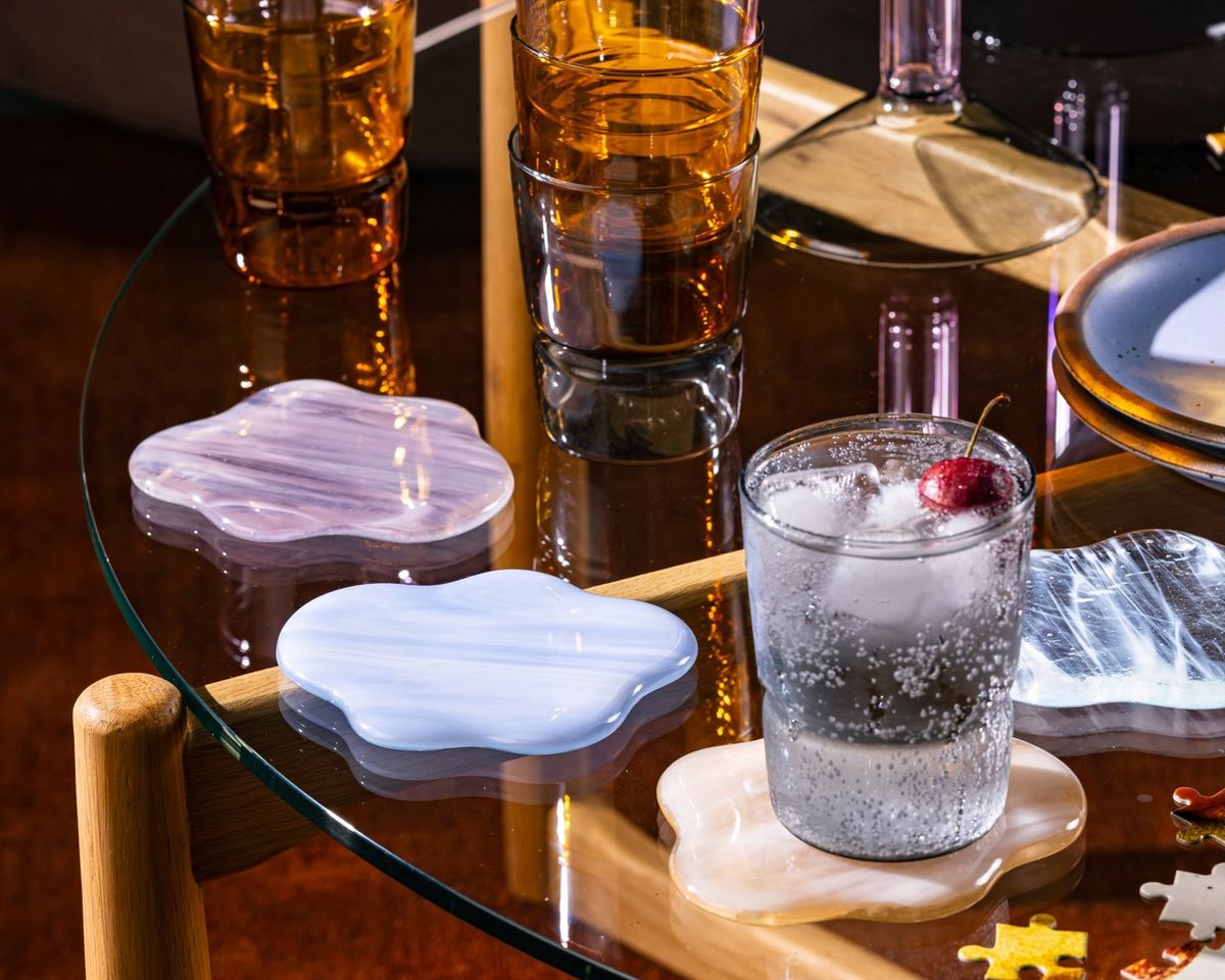 On a table, 4 glass coasters shaped like clouds sit with a simple tumbler filled with a cocktail