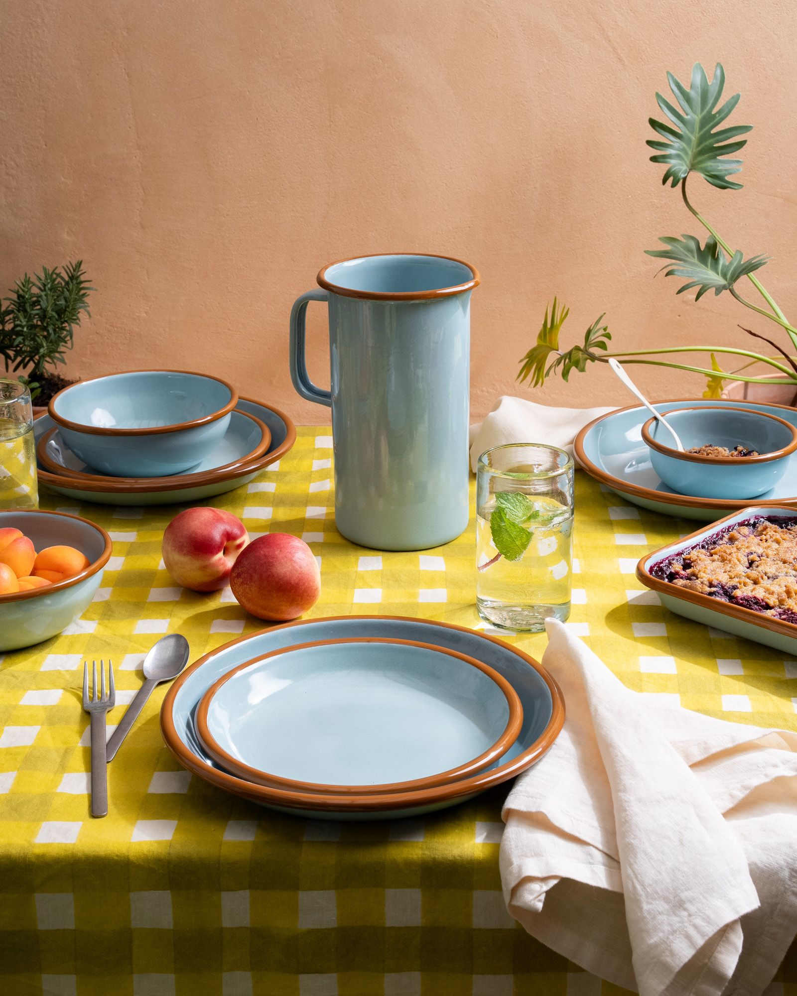 Enamel plates, bowls, and pitchers in a blue color with a brown rim on a yellow crosshatch tablecloth, with other dining accessories.