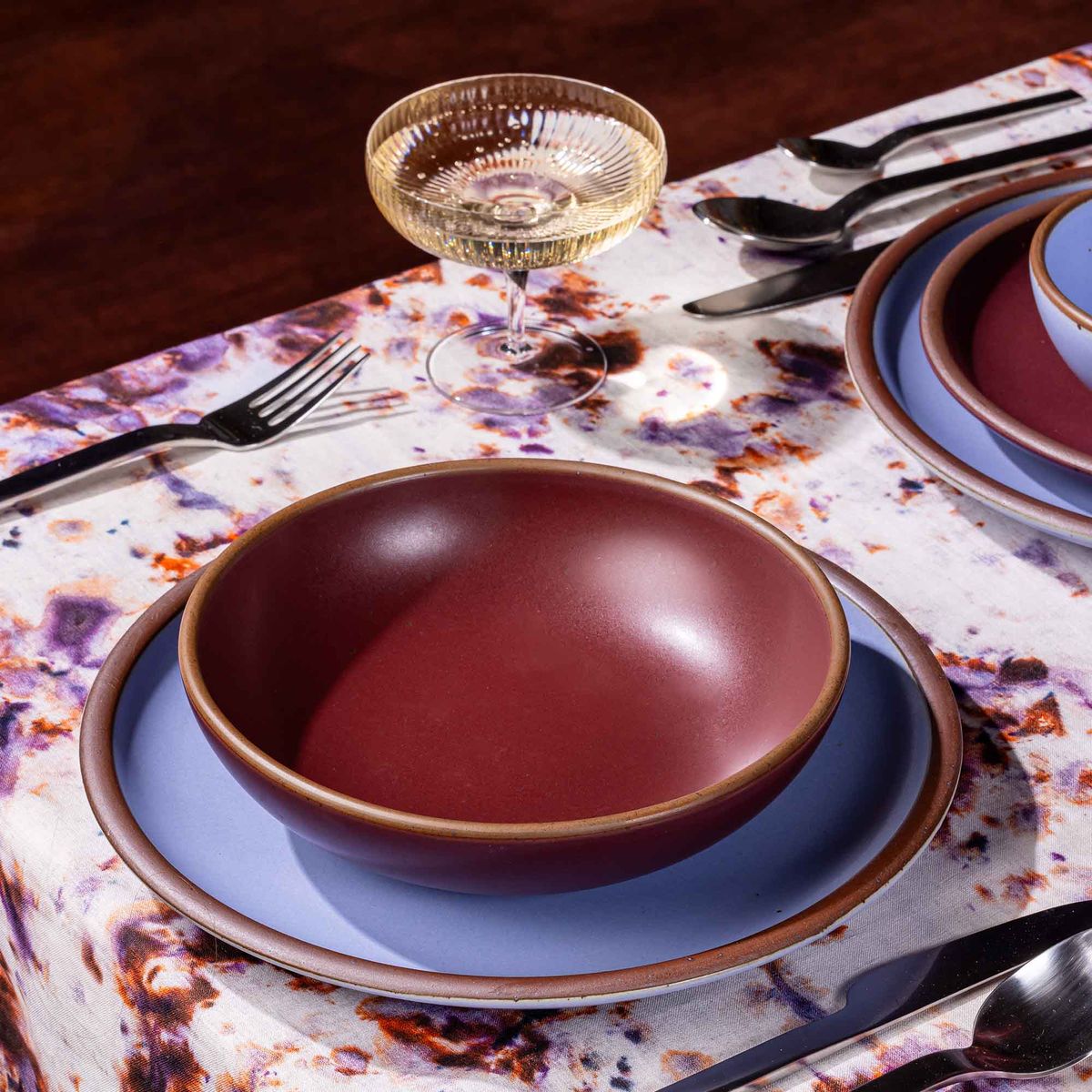 A table setting featuring marbled textiles, a coupe glass, and a plate and shallow bowl in plum and periwinkle colors