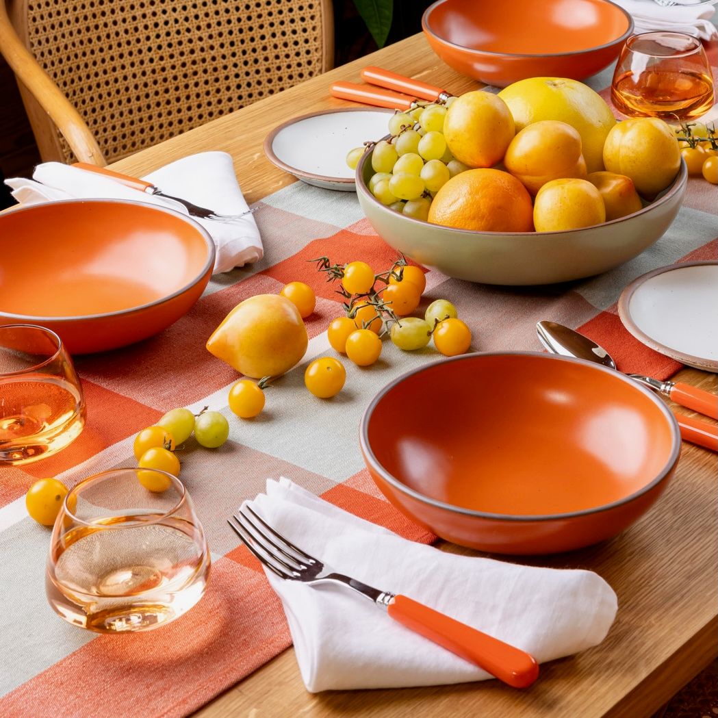 A table filled with several place settings, featuring a shallow dinner bowl in a bold orange, a folded white napkin, orange flatware, whiskey snifters, and a centerpiece filled with fruits and vegetables, all in shades of yellow.